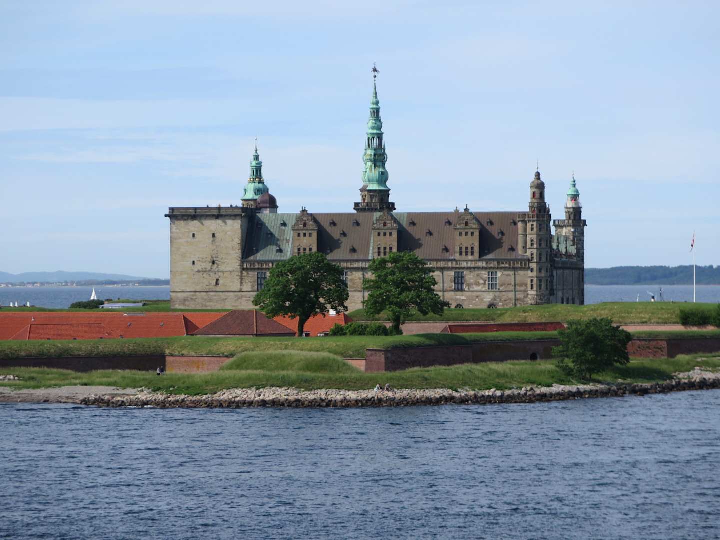 A grand castle-like structure with green-roofed towers stands on a small island surrounded by water, with a grassy foreground and a body of water in the background.