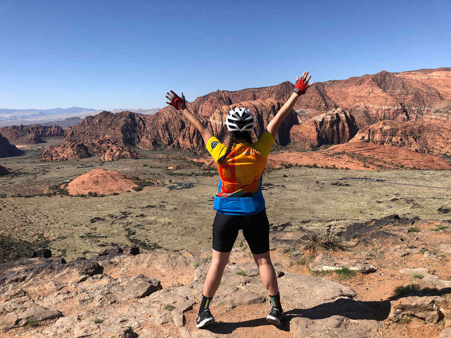 A person in colorful athletic clothing stands with arms raised, surrounded by a dramatic desert landscape of rugged mountains and rocky terrain.
