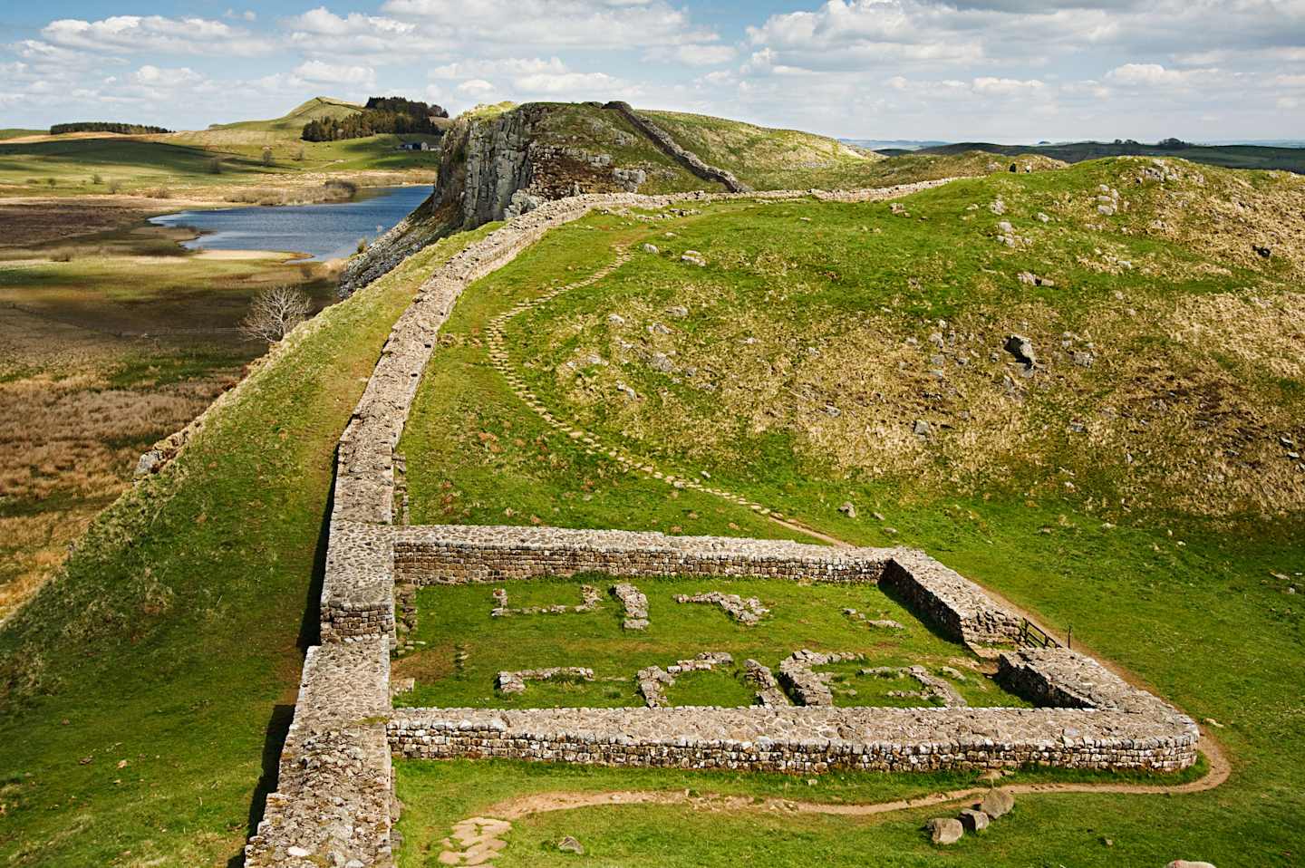 A grassy, hilly landscape with a winding stone path leading to the ruins of an ancient structure, set against a backdrop of cloudy skies and rolling hills.