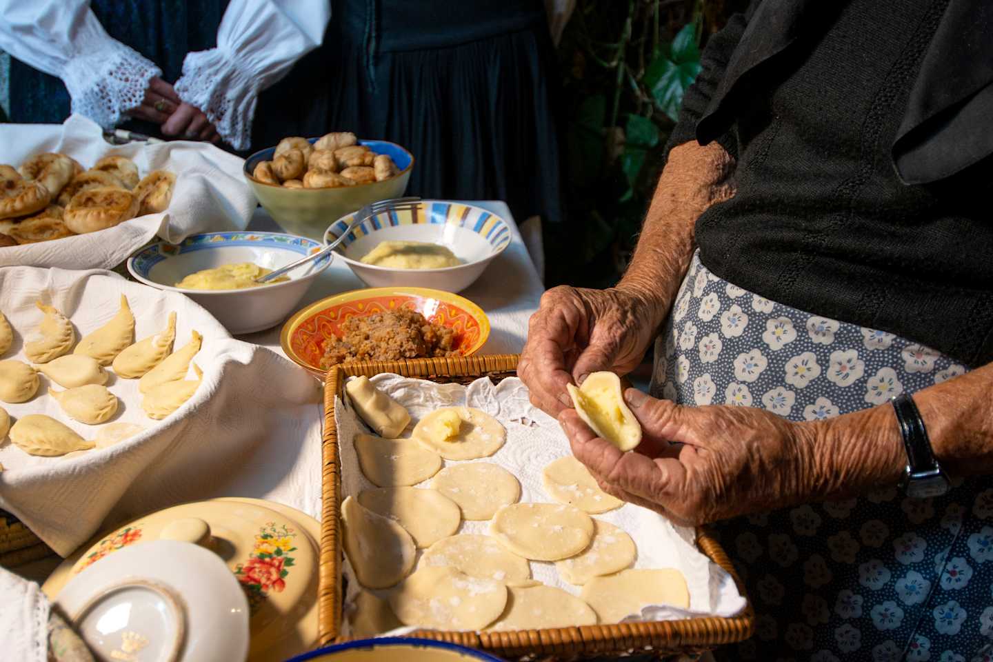 The image depicts a table filled with various traditional food items, including freshly baked pastries, bowls of savory dishes, and a person's hands engaged in the preparation of what appears to be dough or pastry. The background suggests a cozy, domestic setting, with a patterned fabric or clothing visible.