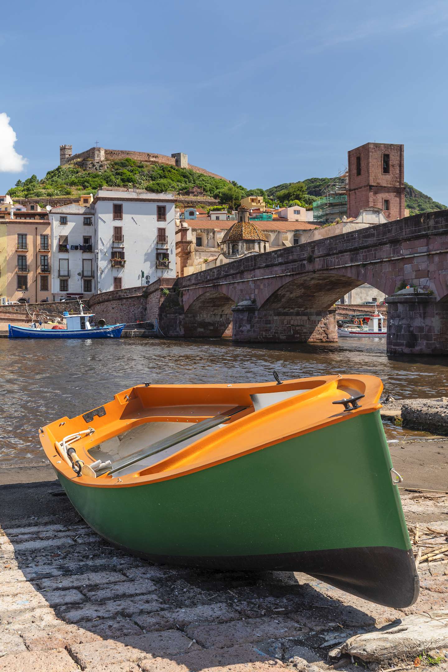 A small green and orange boat rests on the shore, with a historic town and castle visible in the background against a clear blue sky.