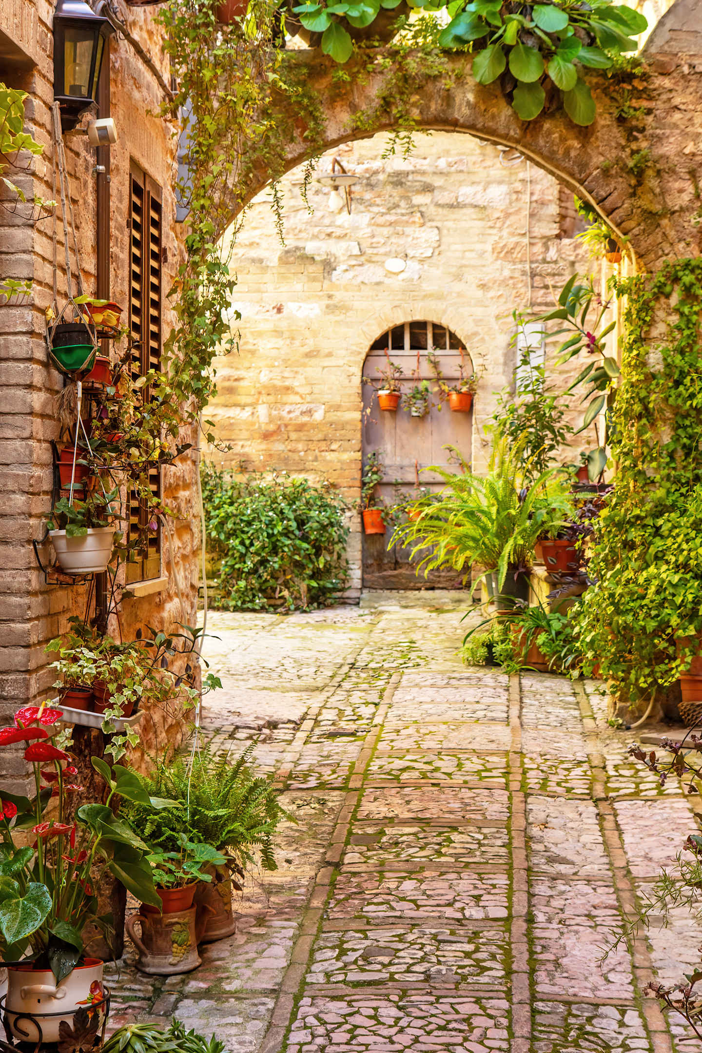A charming, cobblestone courtyard surrounded by lush greenery and a stone archway, creating a cozy and inviting atmosphere.