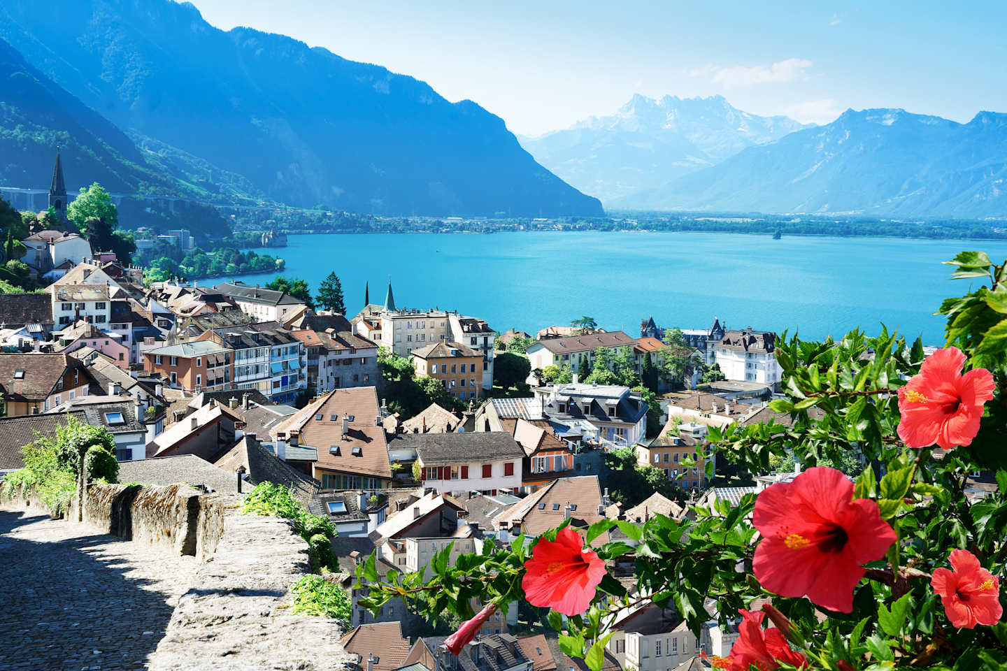 A picturesque town nestled along the shores of a serene lake, surrounded by majestic mountains and adorned with vibrant red hibiscus flowers in the foreground.