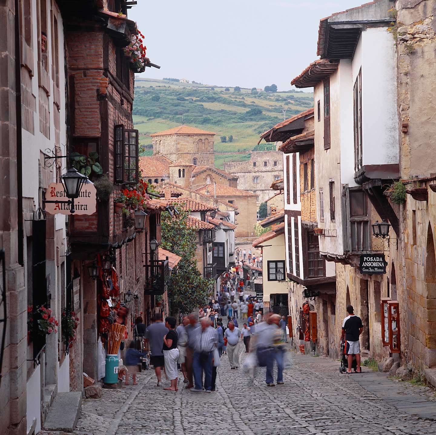 A bustling cobblestone street lined with traditional buildings, shops, and pedestrians, set against a backdrop of rolling hills in the distance.