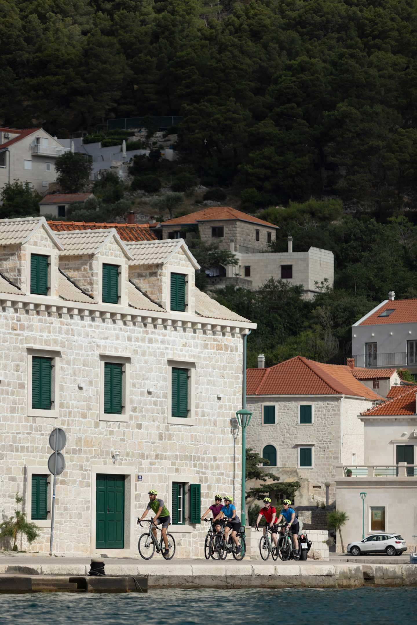 A group of cyclists riding along a coastal town with traditional Mediterranean-style buildings and lush greenery in the background.