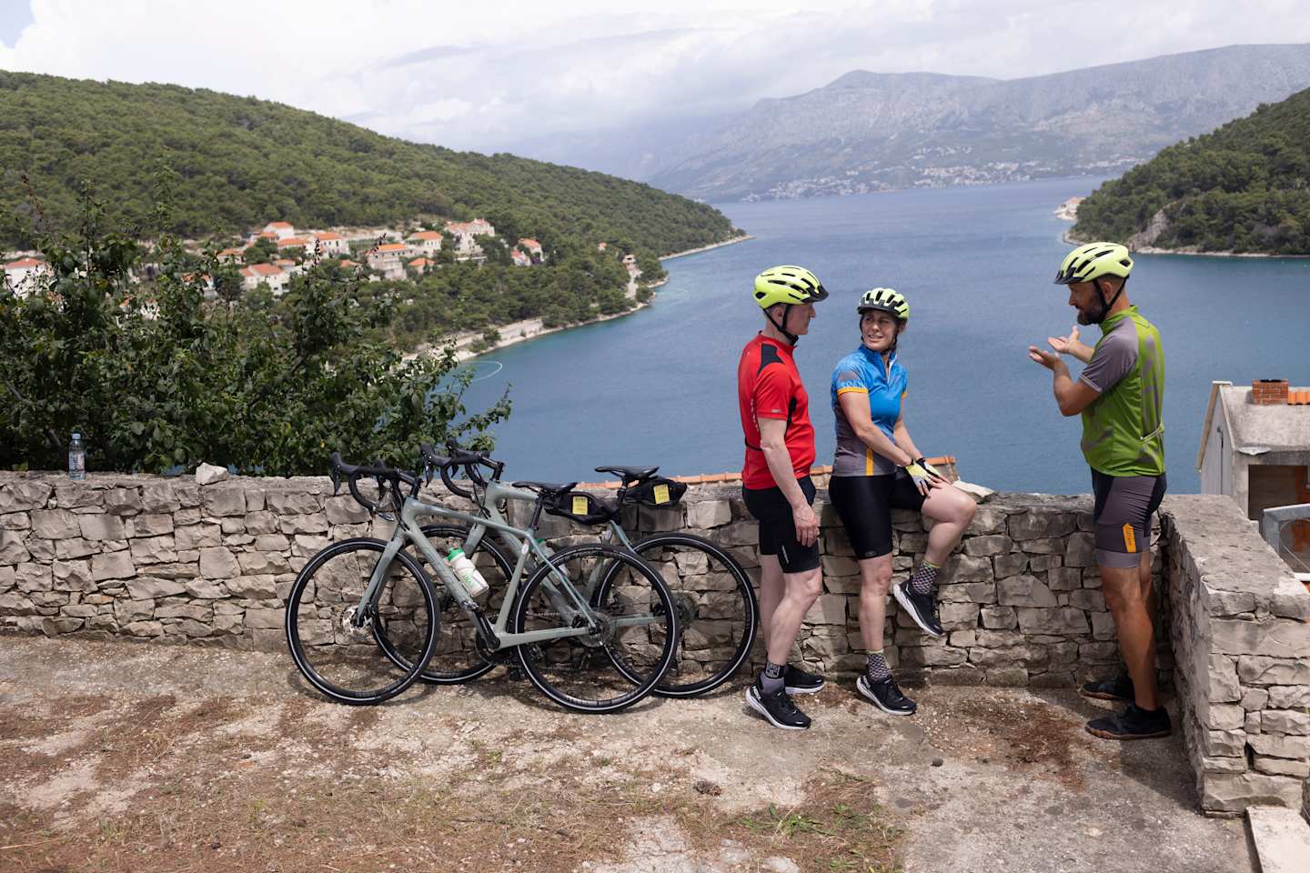 Three cyclists in cycling gear stand next to their bicycles, overlooking a scenic lake surrounded by lush, green hills in the background.