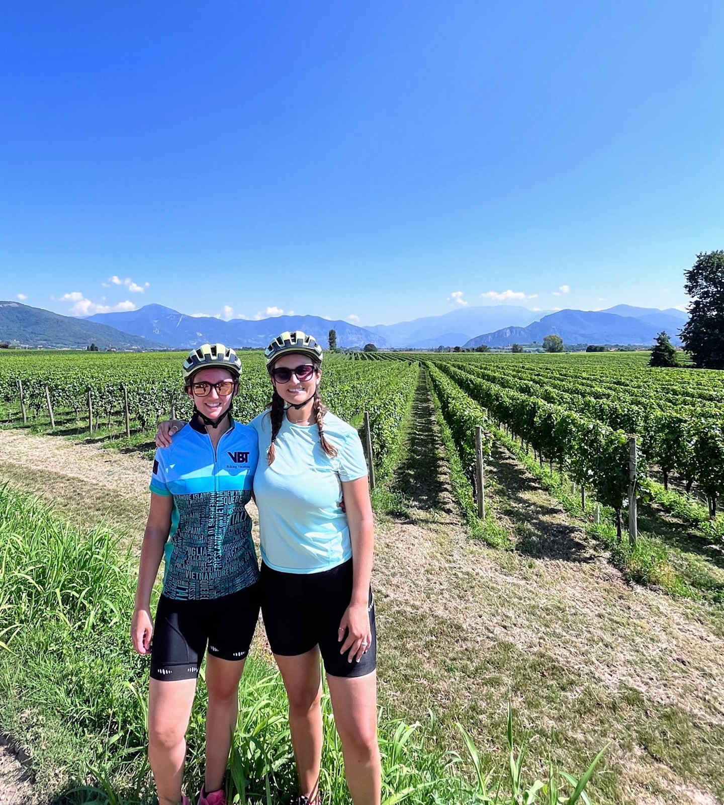 The image shows two women wearing cycling gear standing in a vineyard with mountains in the background.