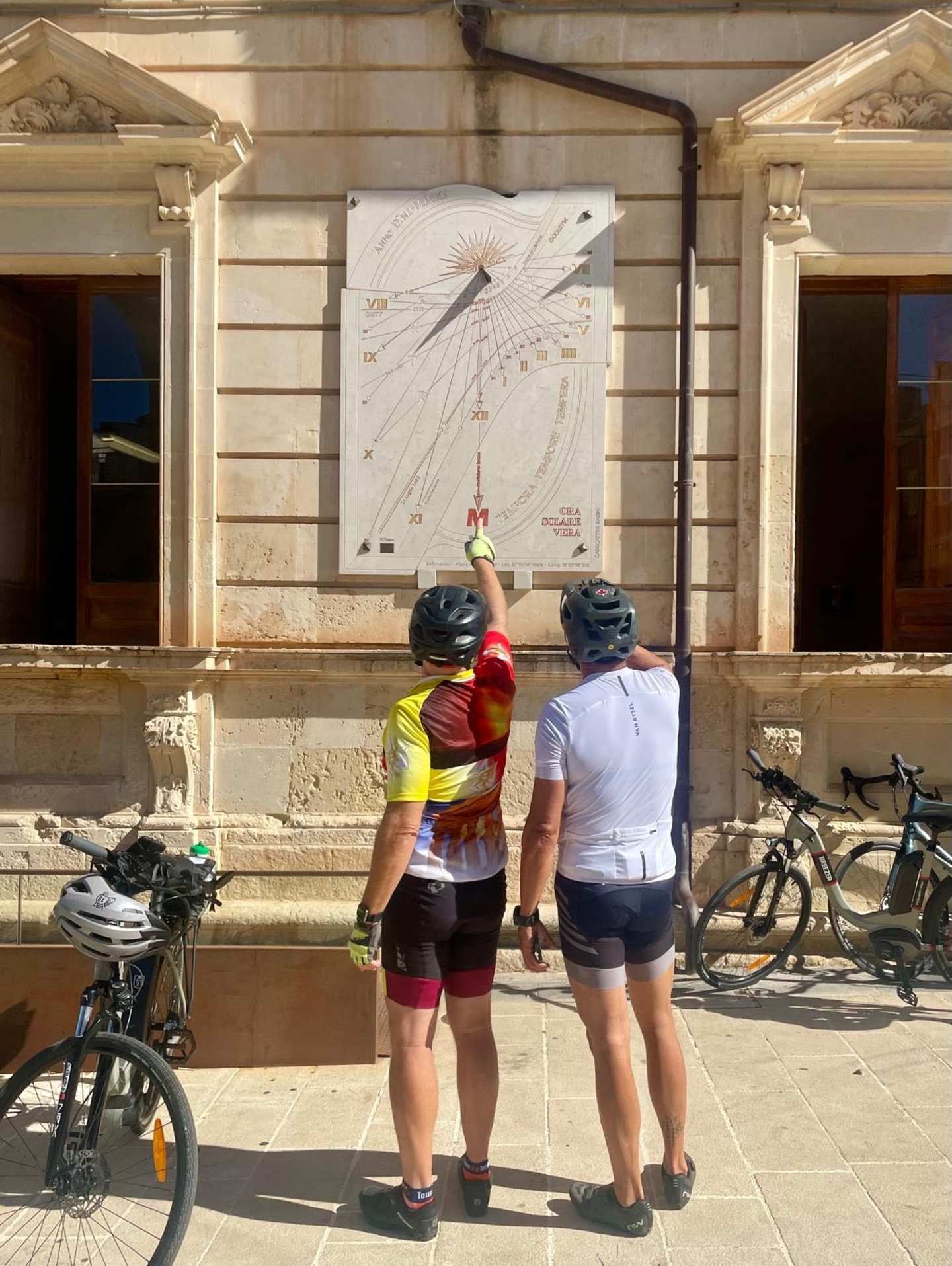 Two individuals in cycling attire are standing in front of a building with ornate architectural features, examining what appears to be a sundial or other outdoor timepiece mounted on the wall.