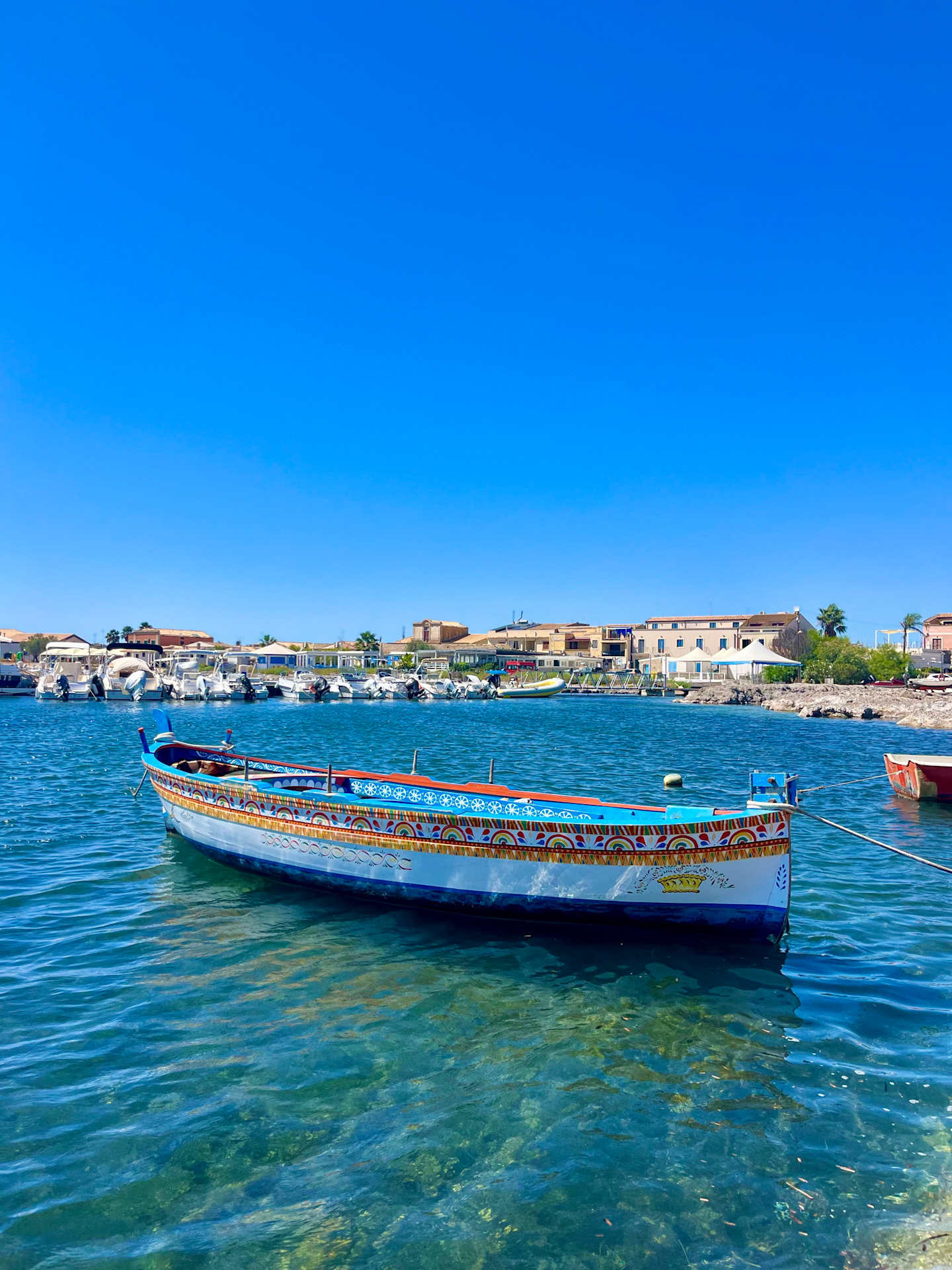 A colorful traditional wooden boat floats on the clear, turquoise waters of a harbor, with a backdrop of buildings and structures along the shoreline under a vibrant blue sky.