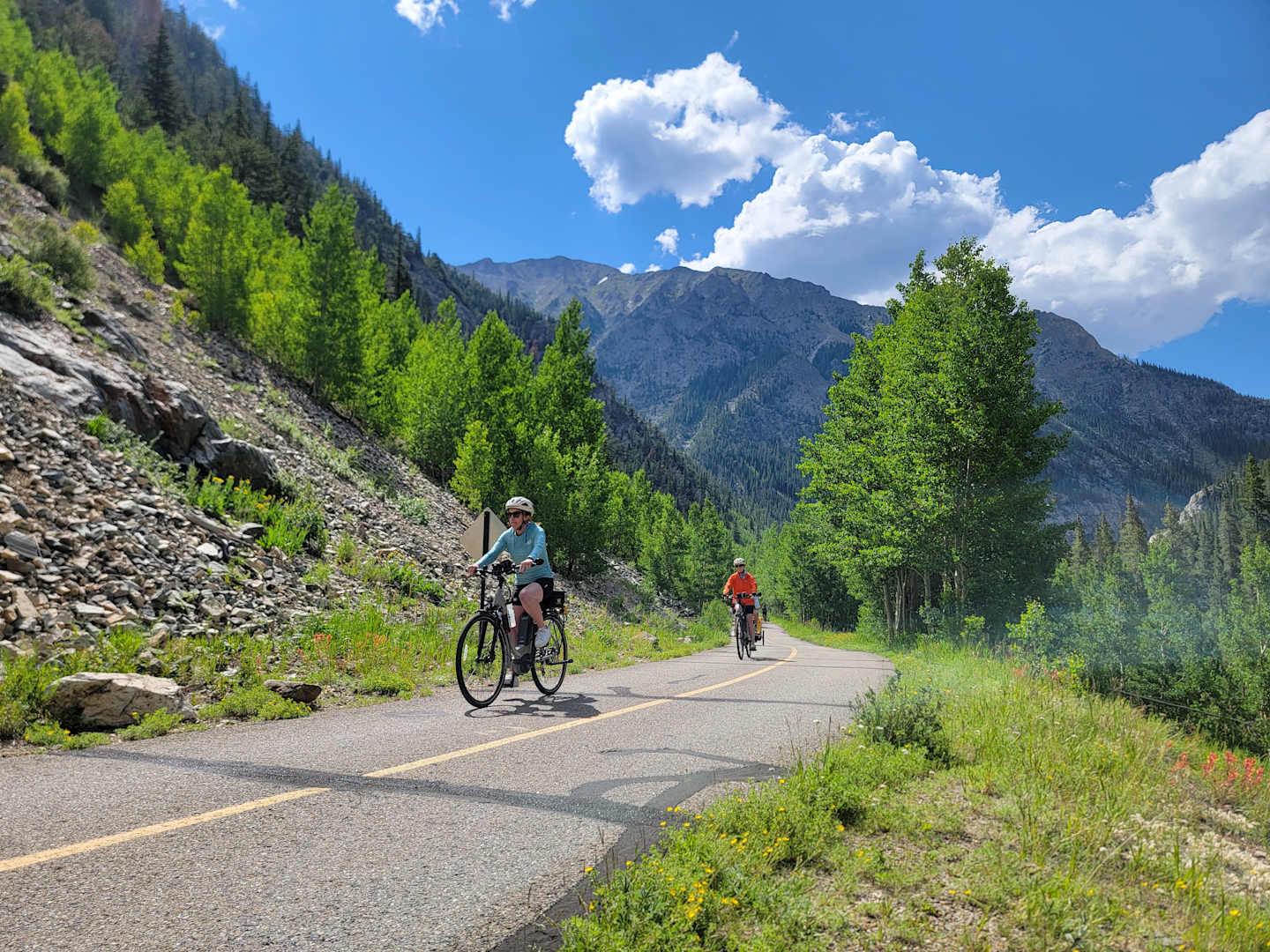 A scenic mountain road with cyclists riding through a lush, forested landscape under a blue sky with fluffy clouds.