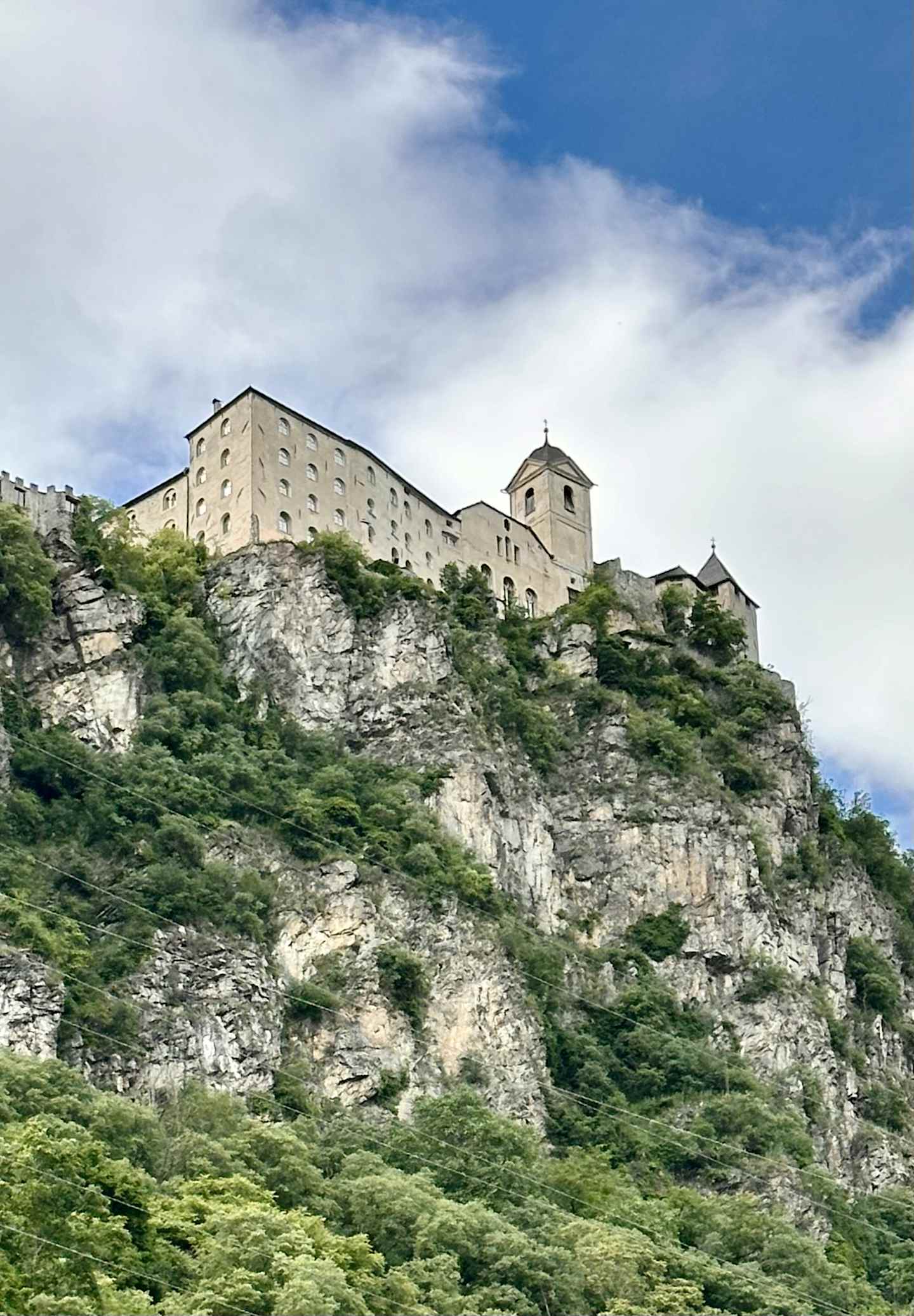 A large, imposing castle or fortress sits atop a steep, rocky cliff, surrounded by lush, green vegetation in the foreground.