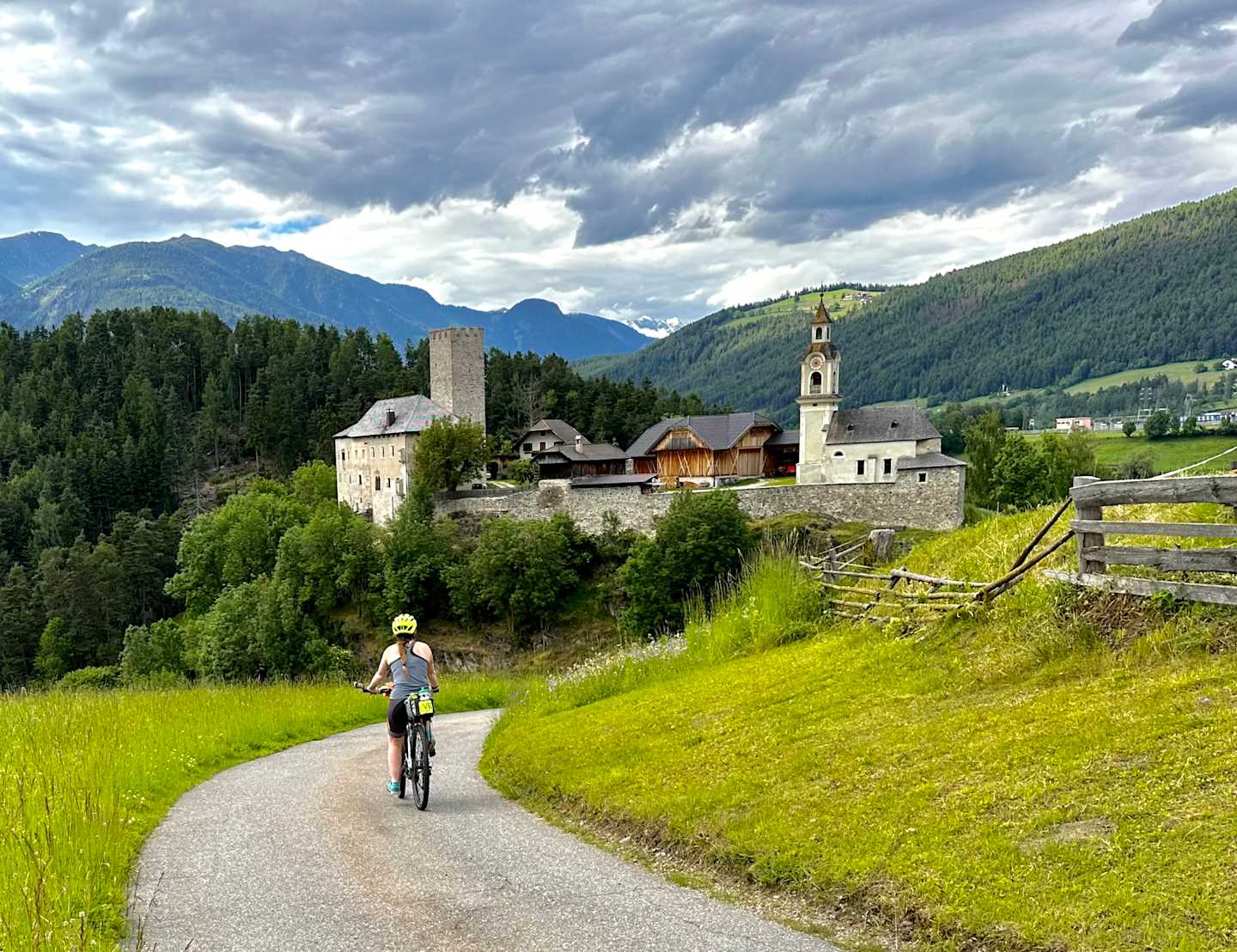 A person on a bicycle rides along a dirt path through a lush, green meadow, with a picturesque medieval castle nestled among the mountains in the background under a dramatic cloudy sky.