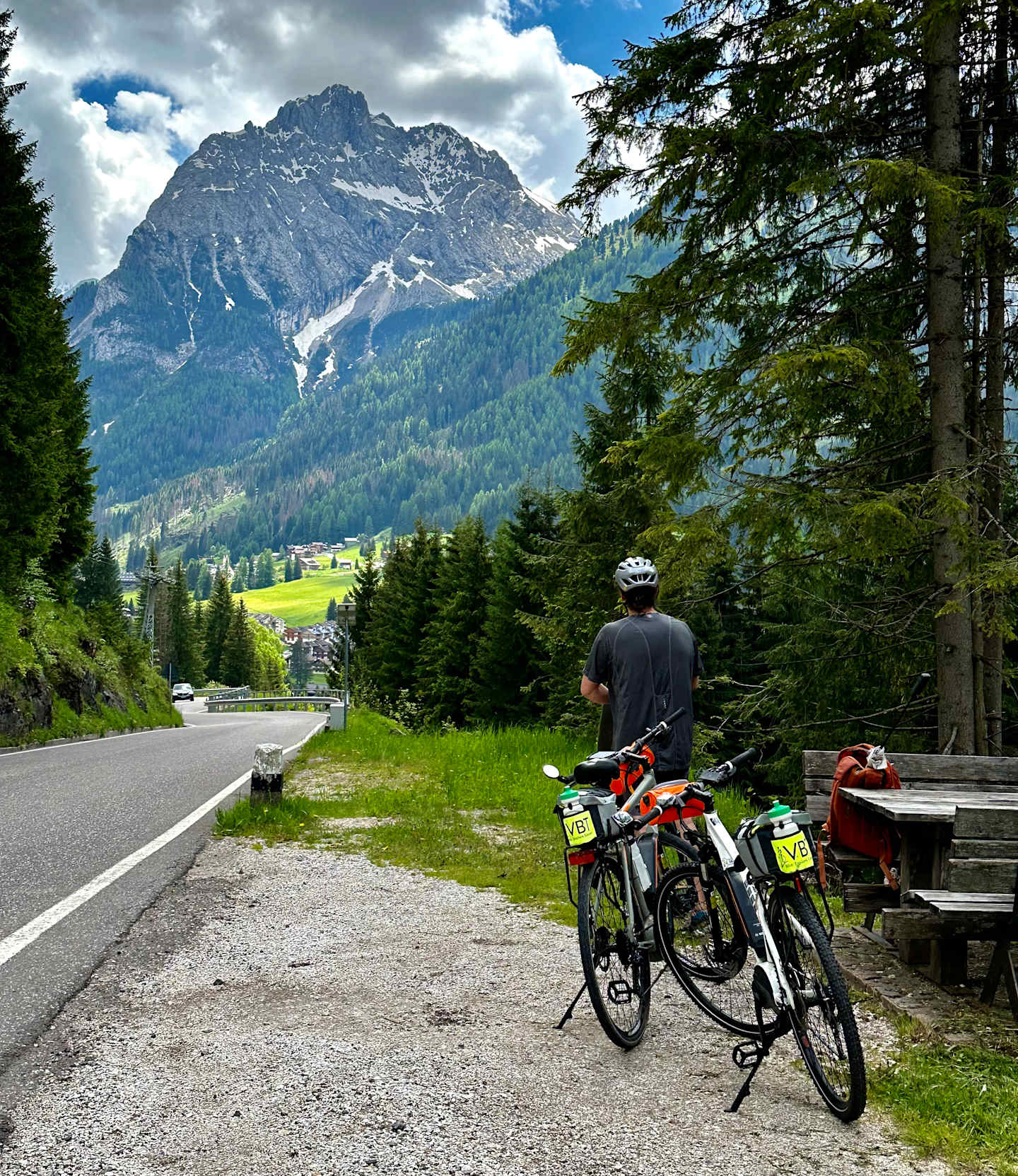 A cyclist stands on a road surrounded by lush green forests and towering snow-capped mountains in the distance.