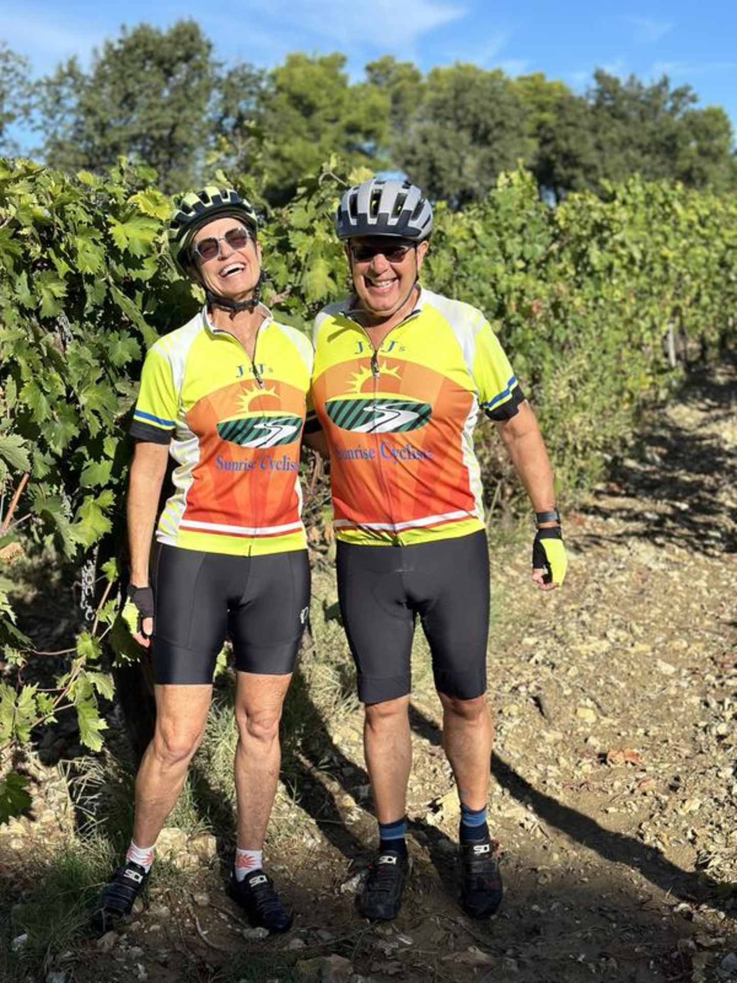 Two cyclists in bright cycling gear stand on a dirt path surrounded by lush greenery and trees.