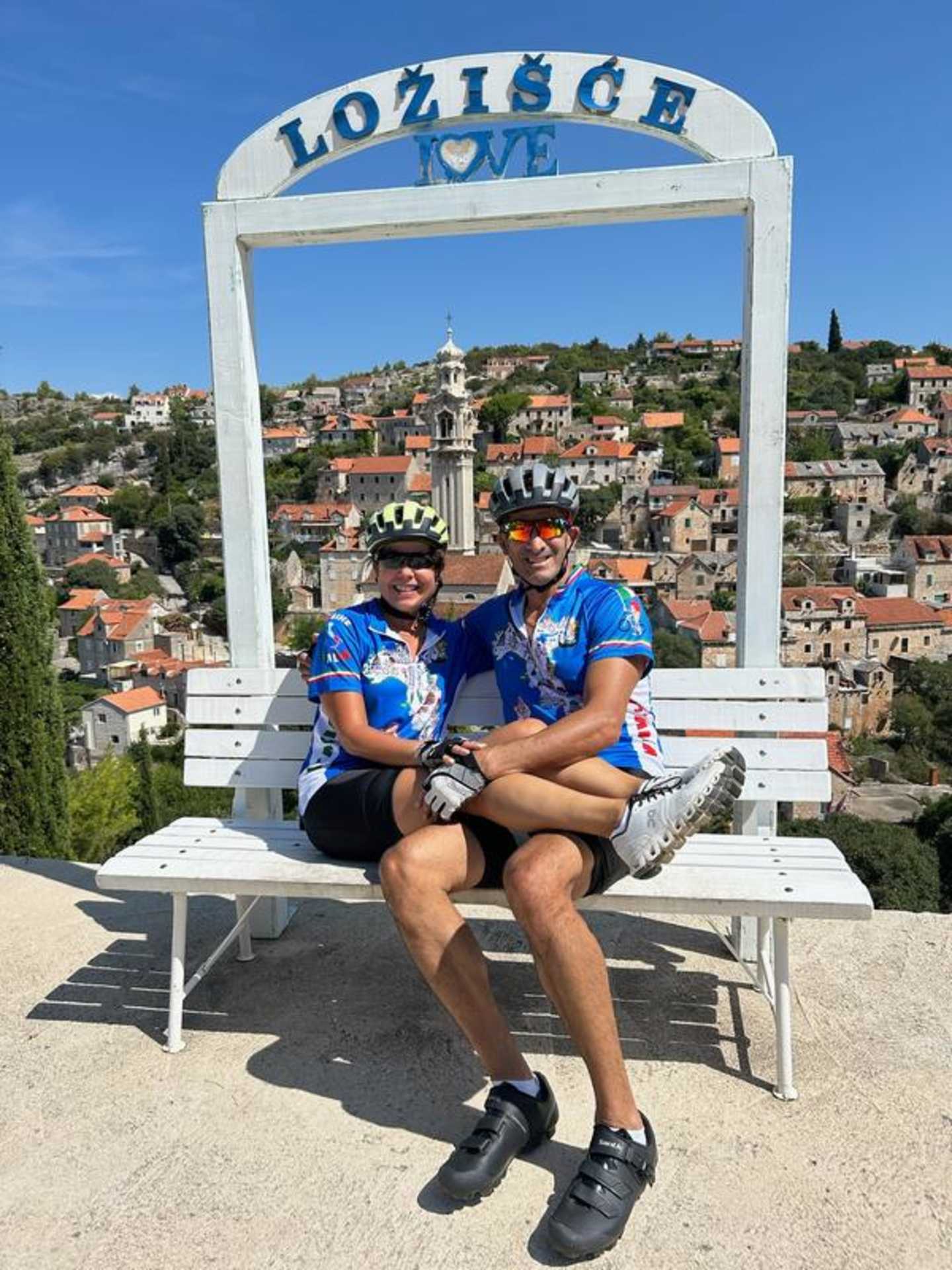 Two people, a man and a woman, are sitting on a bench in the foreground, with a scenic town in the background framed by a large "Lozisce" sign.
