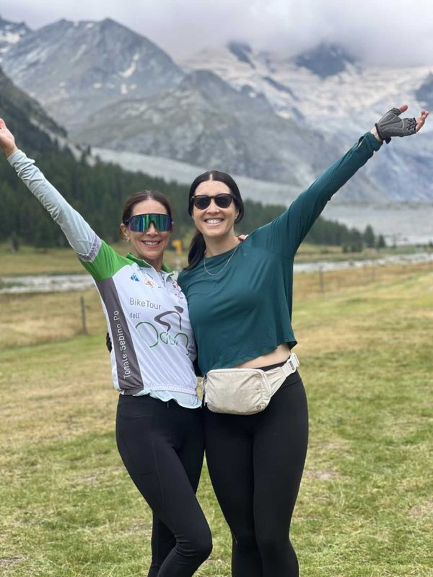 Two women standing in a grassy field with mountains in the background, their arms raised in a celebratory gesture.