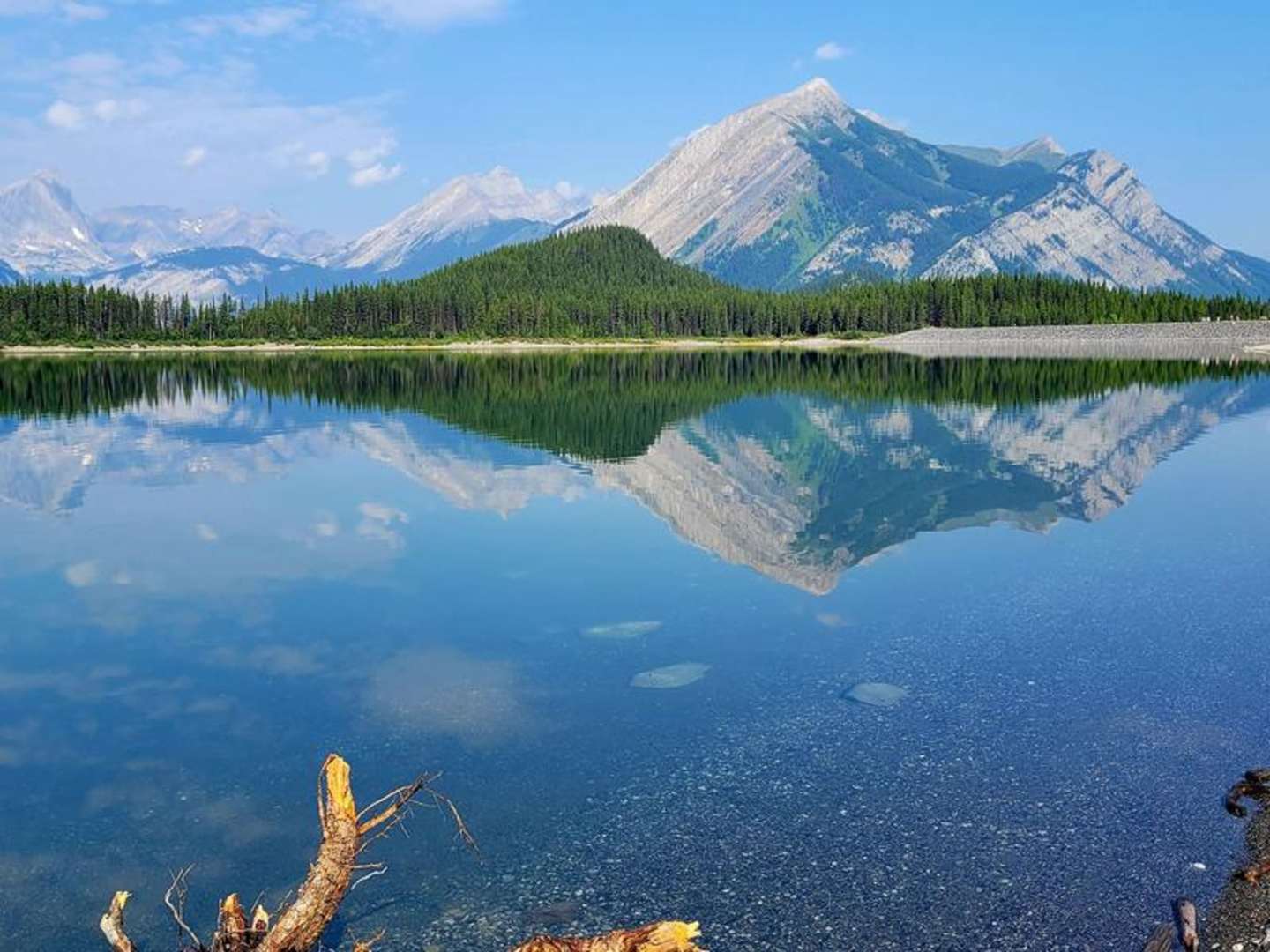 Majestic snow-capped mountains rise in the background, reflected perfectly in the serene, mirror-like lake in the foreground, surrounded by a lush, evergreen forest.