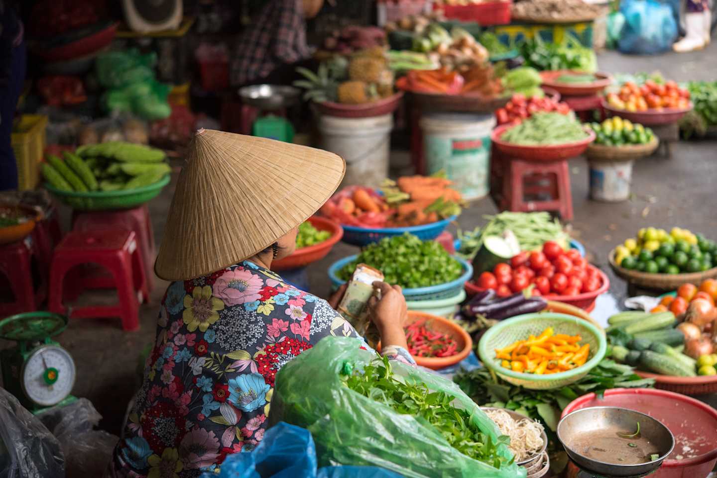 A vibrant and colorful outdoor market stall filled with an array of fresh produce, including various vegetables, fruits, and other food items, with a person wearing a traditional Vietnamese conical hat visible in the foreground.
