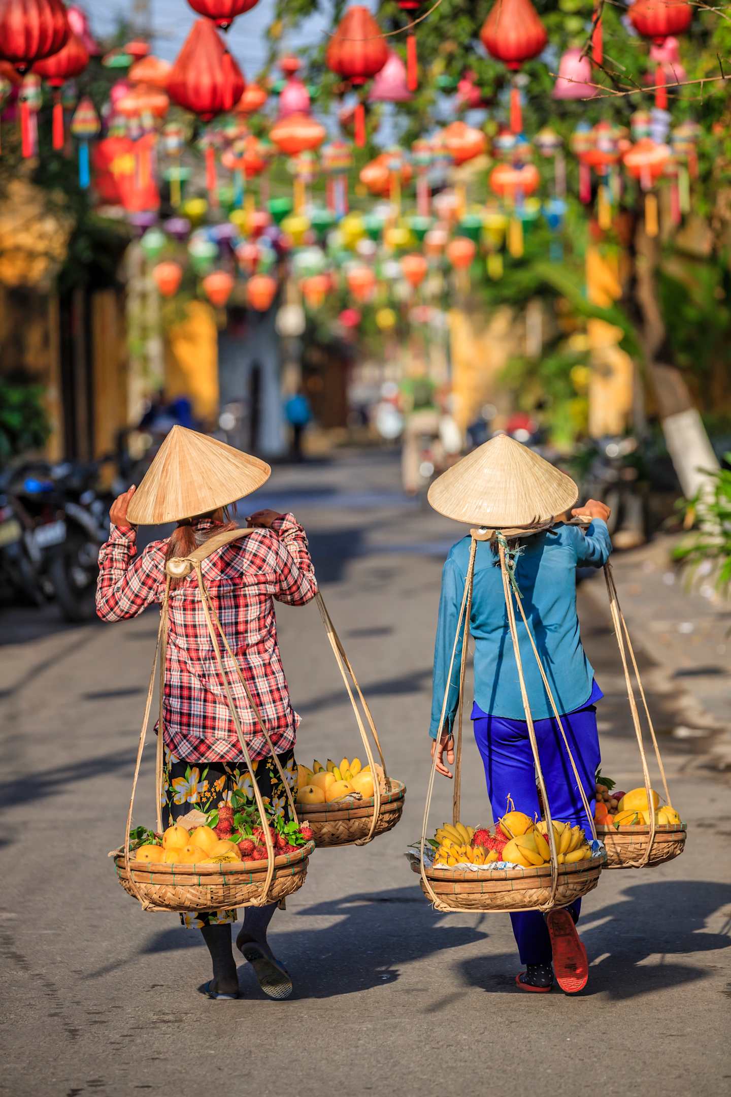 Two people wearing traditional conical hats and carrying baskets of fruits and vegetables on a street lined with colorful lanterns and buildings.