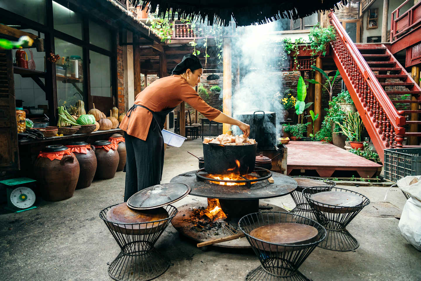 A bustling outdoor kitchen scene with a person cooking over a large fire pit, surrounded by various cooking vessels and a lush, tropical-looking environment.