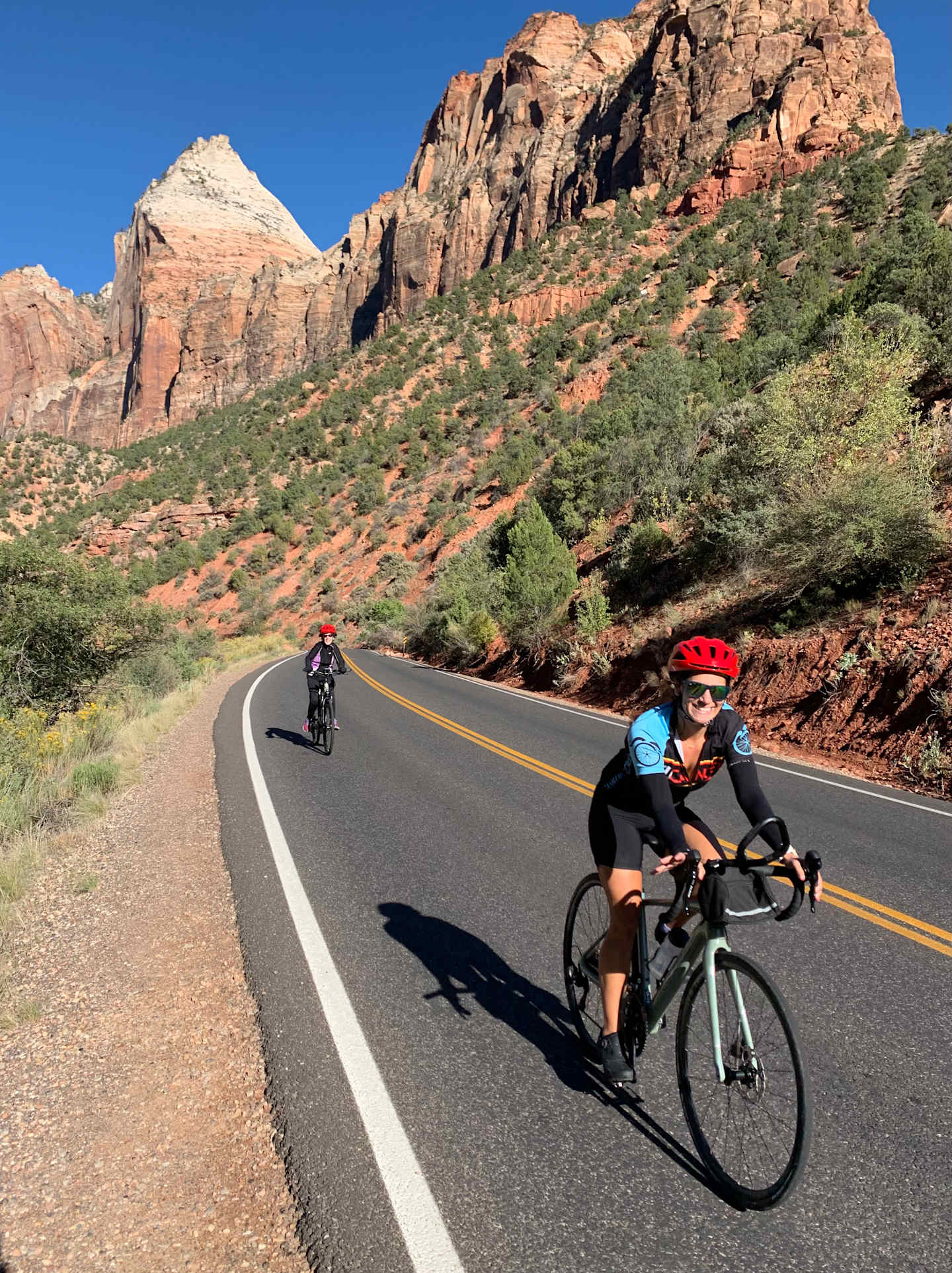 The image depicts a scenic road winding through a rugged, mountainous landscape, with two cyclists riding their bicycles along the road.