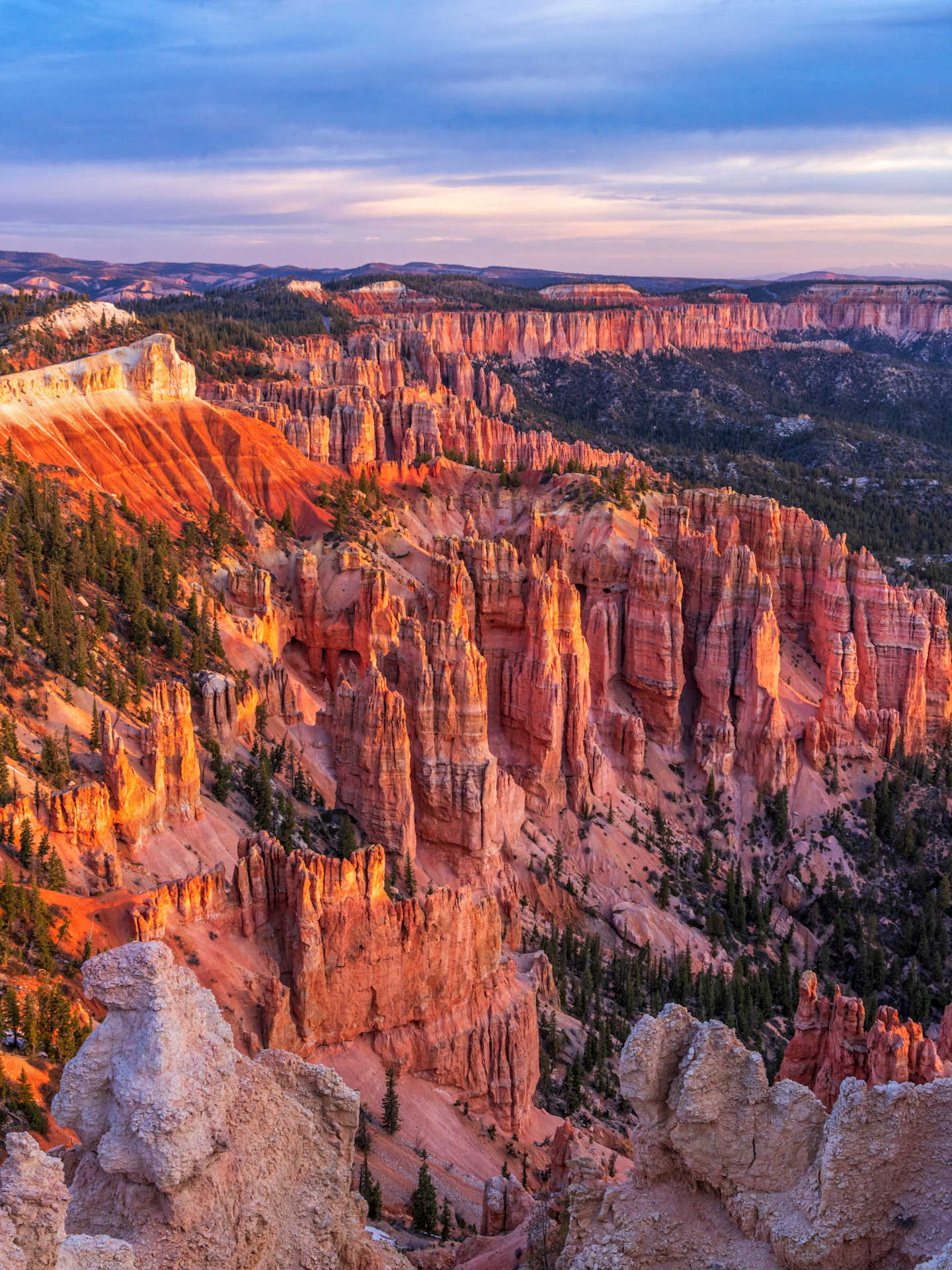A vast, rugged landscape of towering red rock formations and deep canyons, set against a dramatic sky with warm hues of orange and blue.