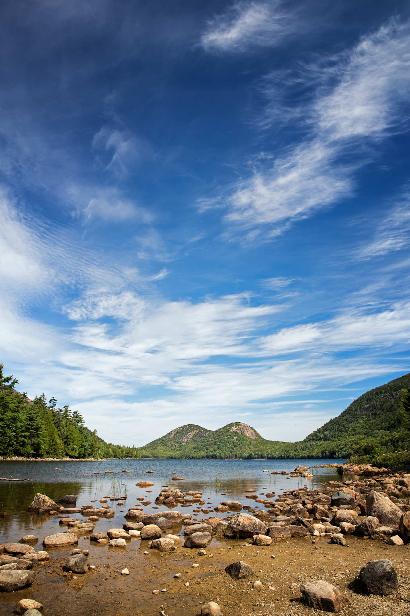 A serene lake surrounded by rocky shores and lush green hills, with a picturesque blue sky dotted with wispy clouds overhead.
