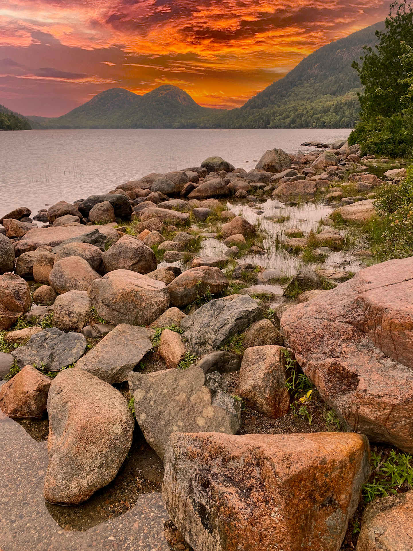 A picturesque landscape with a rocky shoreline leading to a serene lake, surrounded by lush hills and a dramatic, fiery sunset sky.