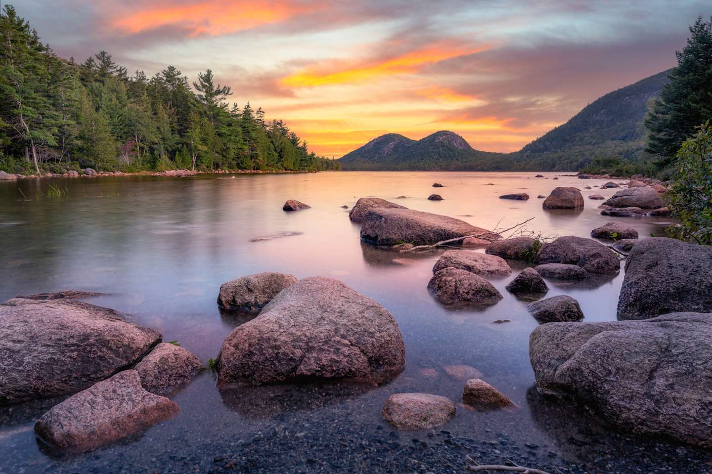 A serene lake surrounded by rocky shores and lush evergreen forests, with a stunning sunset sky casting vibrant hues of orange and pink over the tranquil scene.