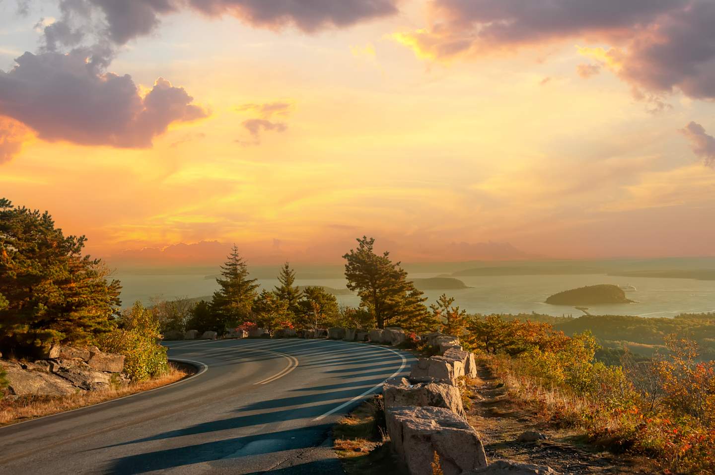 A winding road leads through a scenic landscape, with a vibrant sunset casting a warm glow over the distant islands and the surrounding forest.