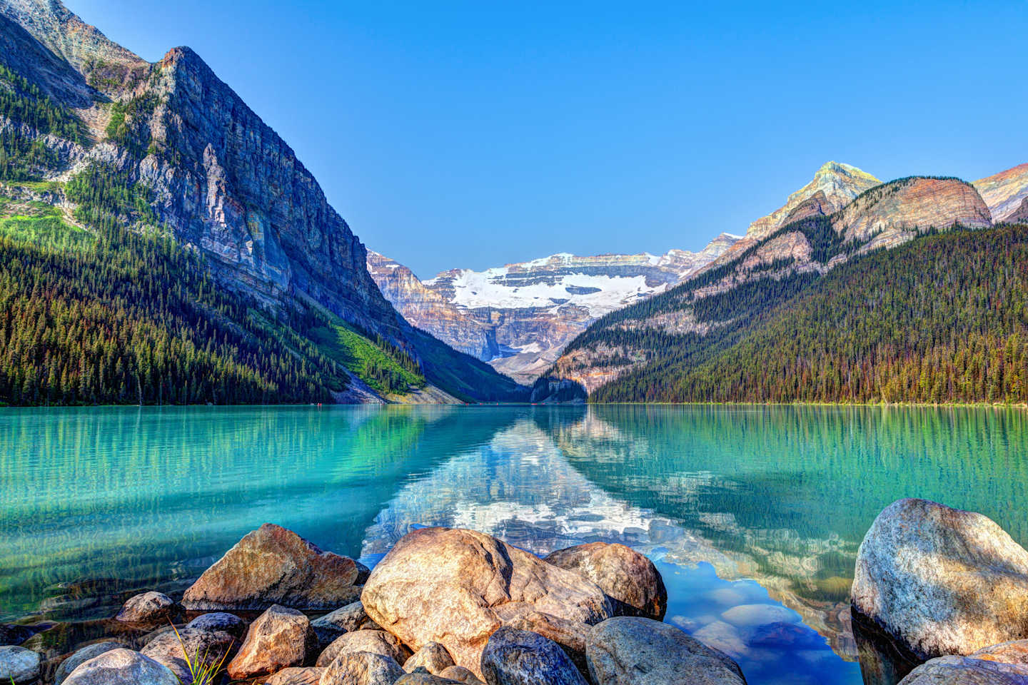 A serene, turquoise lake surrounded by majestic, snow-capped mountains and lush, forested slopes, with large boulders in the foreground reflecting the stunning natural scenery.