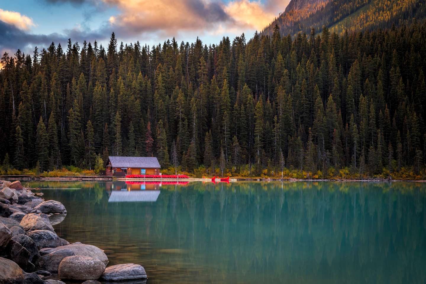 A serene mountain lake surrounded by a dense forest, with a small red cabin reflected in the calm, turquoise waters, set against a backdrop of dramatic, cloud-filled skies.