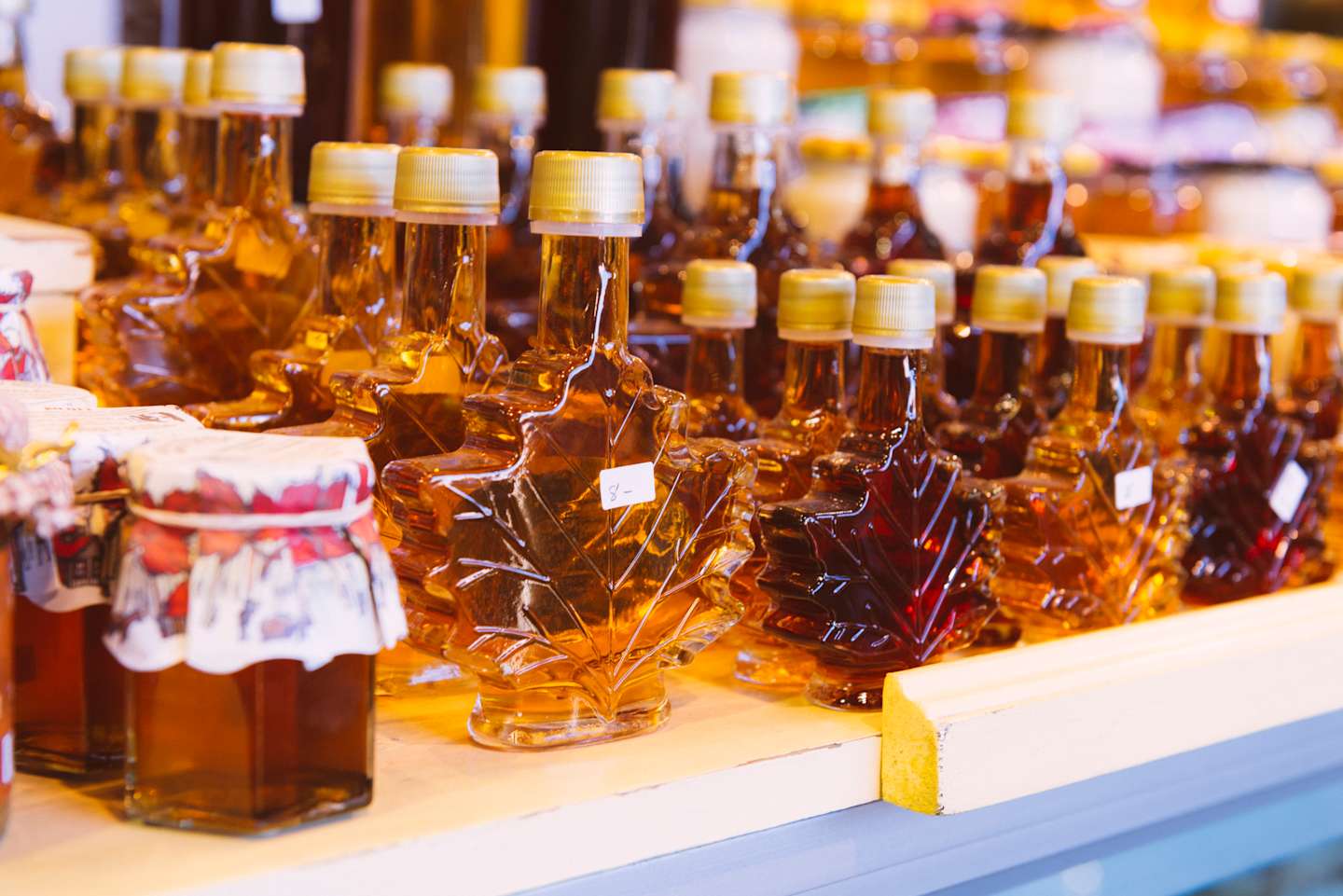 The image depicts a display of various maple syrup bottles in different shapes and sizes, arranged on a shelf against a blurred background.