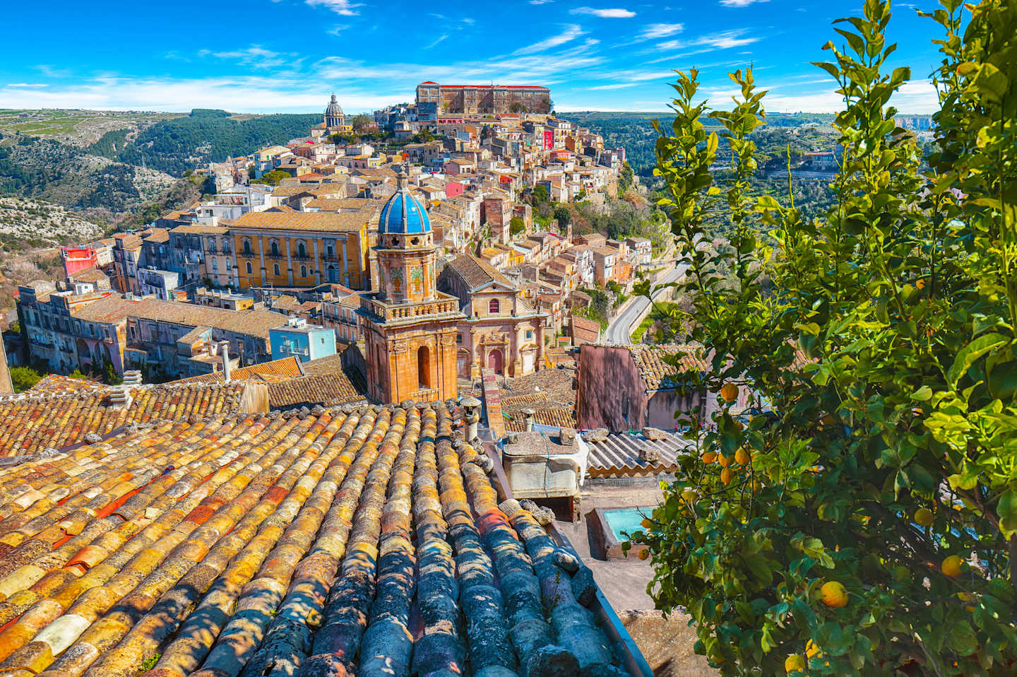 A vibrant and picturesque town nestled in a hilly landscape, with colorful buildings, terracotta roofs, and lush greenery in the foreground, set against a backdrop of a clear blue sky.
