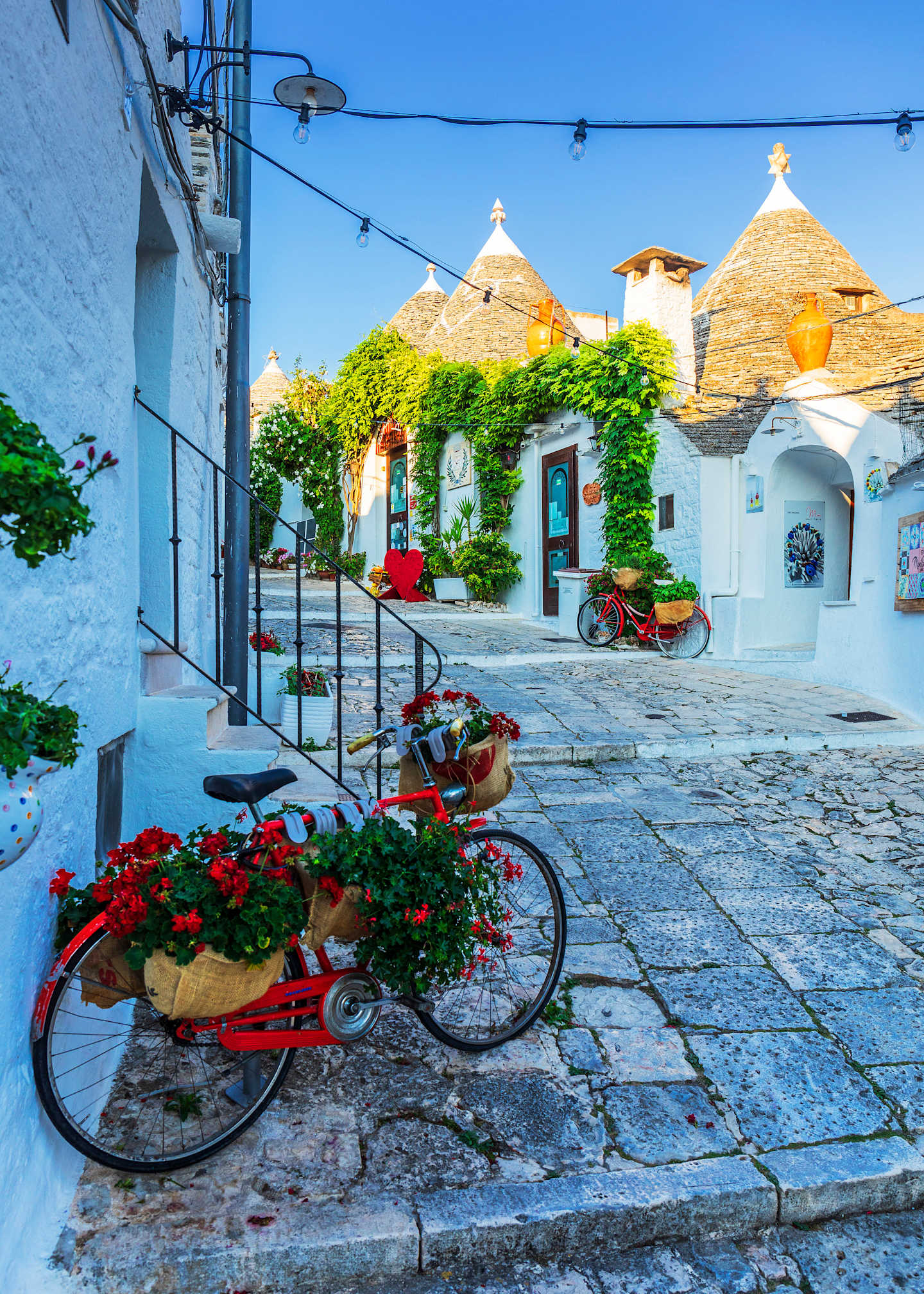 A charming cobblestone street lined with colorful buildings, flower-adorned bicycles, and distinctive conical roofs, set against a vibrant blue sky.