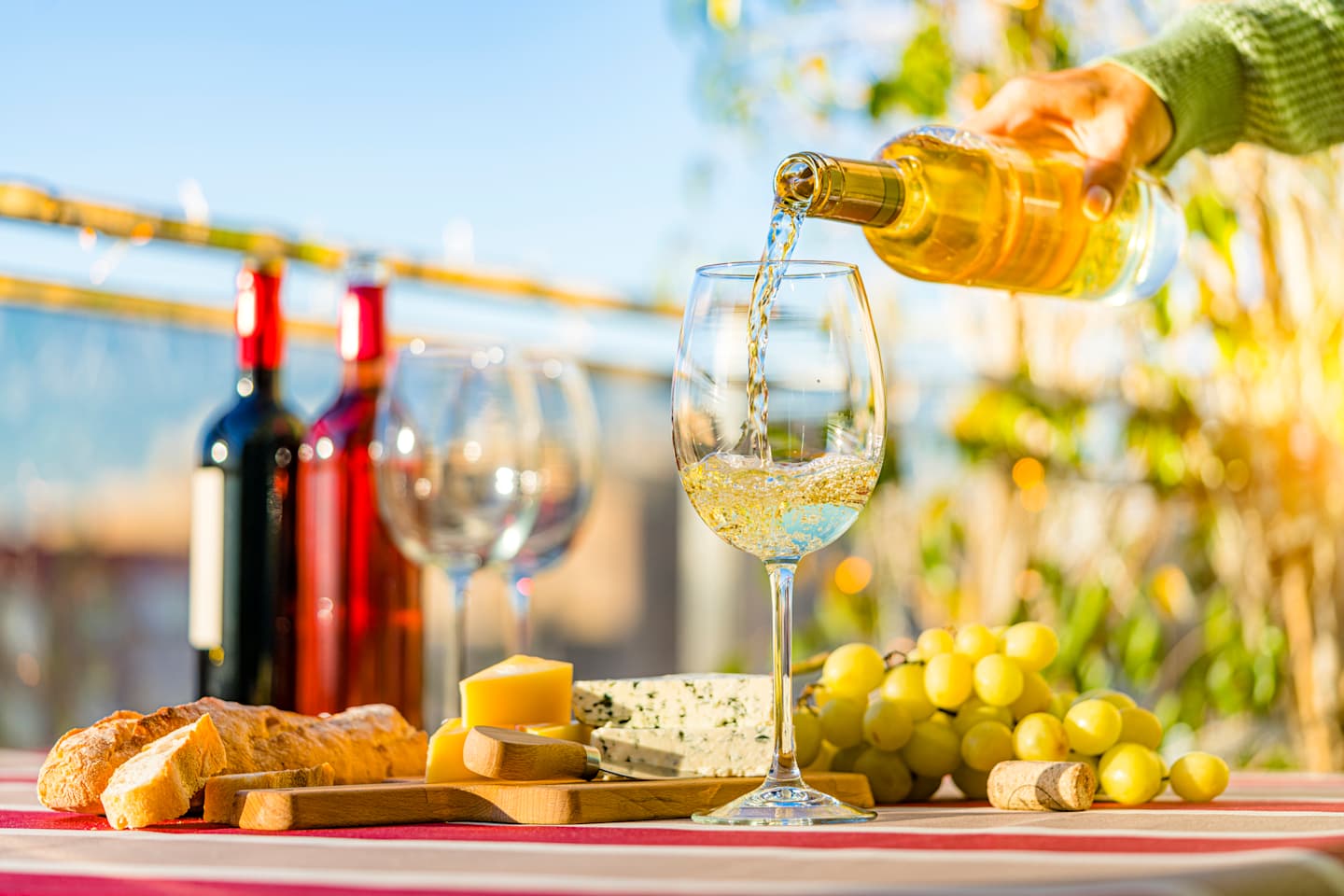 A glass of white wine being poured, surrounded by various cheeses, grapes, and a bottle of red wine, set against a backdrop of a sunny outdoor scene with greenery.