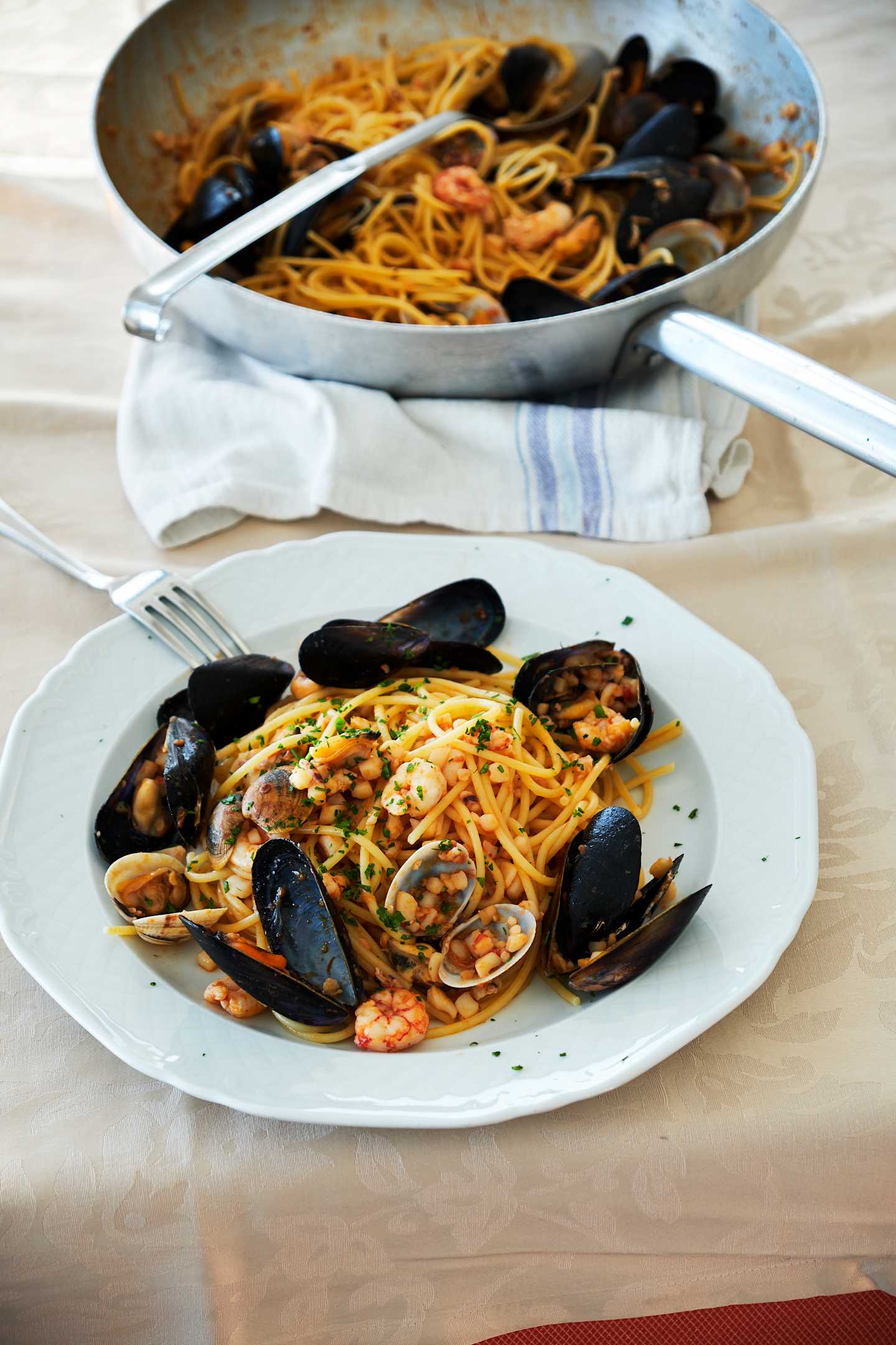 A plate of spaghetti with mussels, garnished with parsley, and a larger serving dish containing the same dish in the background, resting on a white cloth.