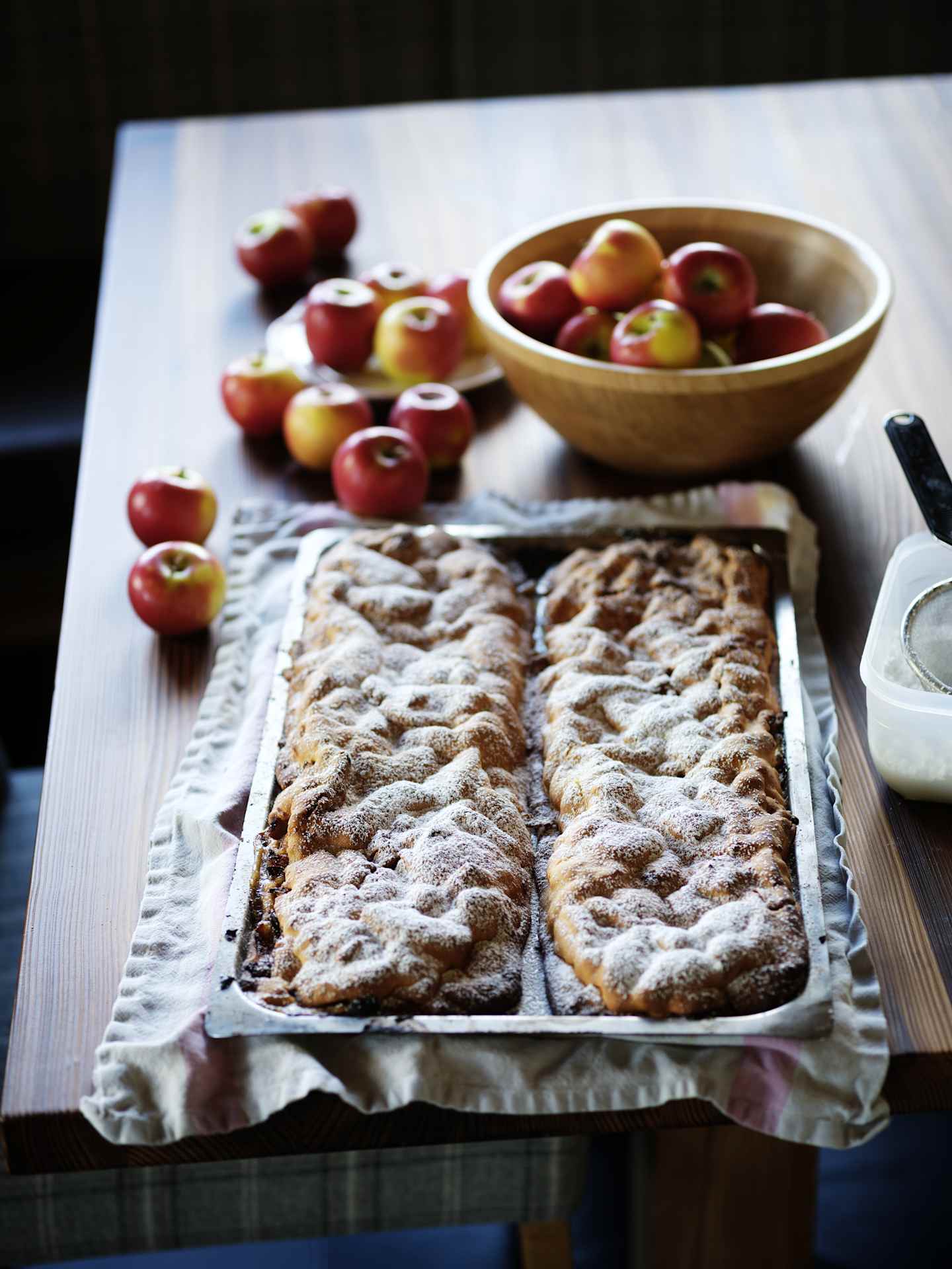 A wooden tray holds a freshly baked apple pie or tart, accompanied by a bowl of ripe, red apples on a wooden surface.