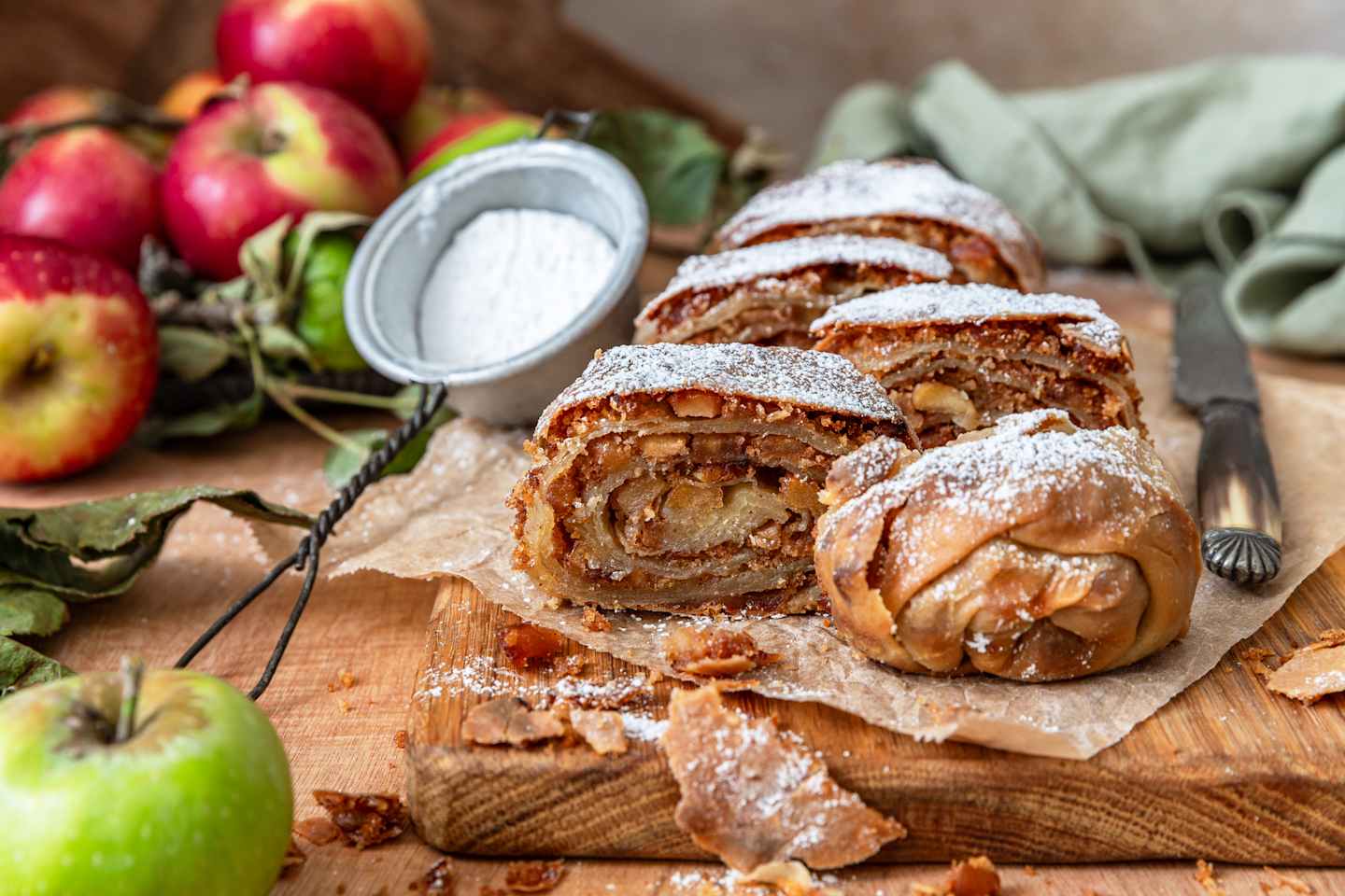 A delicious-looking apple strudel or pastry roll, dusted with powdered sugar, sits on a wooden surface surrounded by fresh green apples.