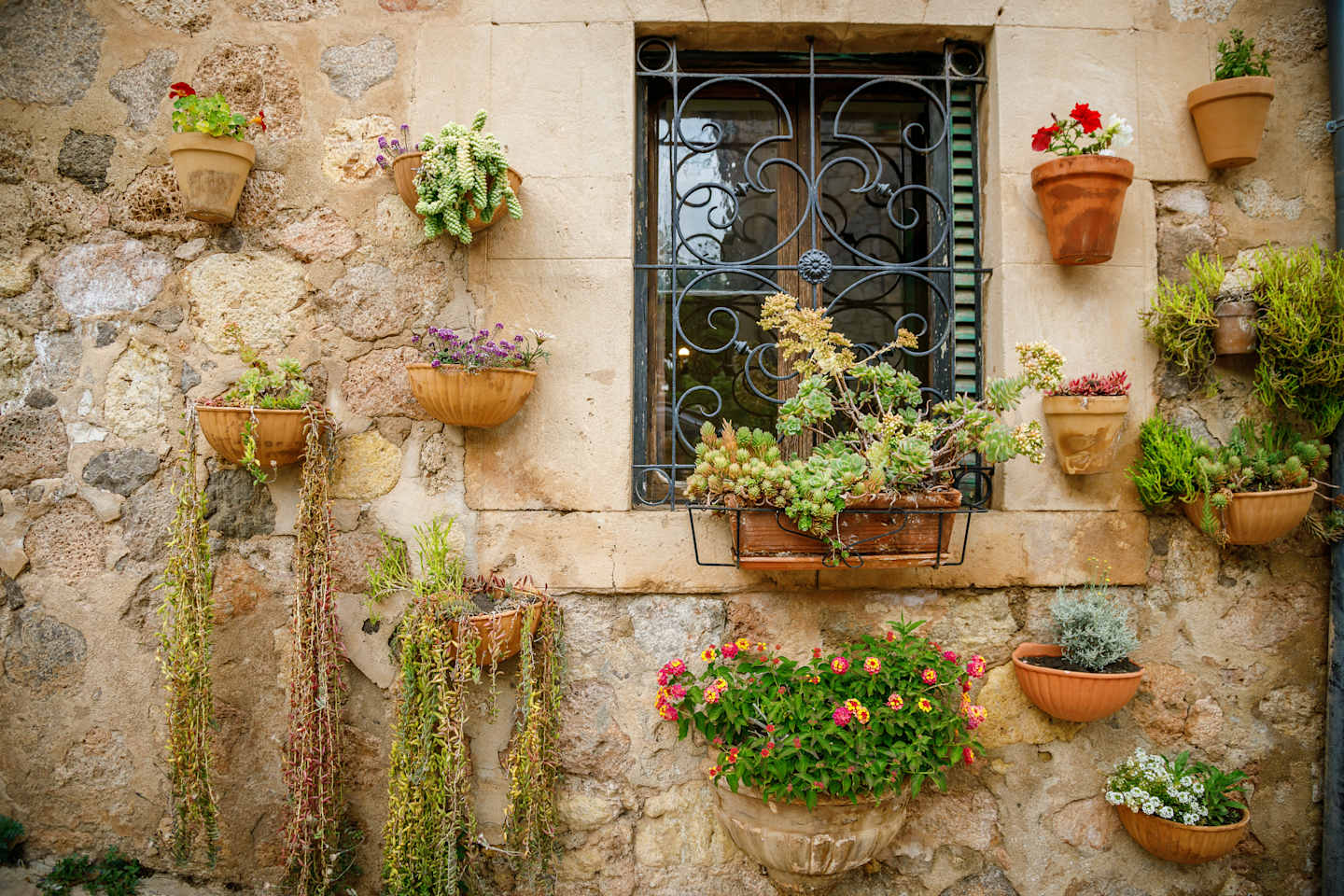 A charming stone wall adorned with an array of potted plants, hanging baskets, and a decorative wrought-iron window frame, creating a vibrant and inviting outdoor scene.