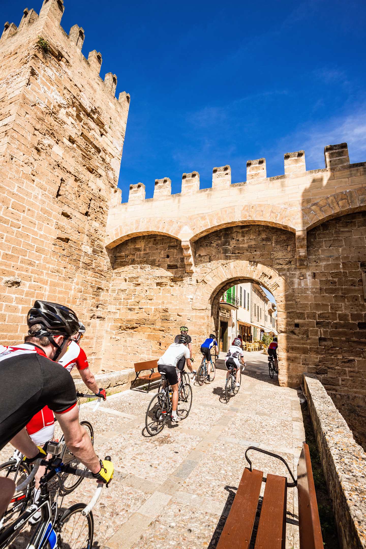 A group of cyclists riding through an archway in a historic stone castle, with a clear blue sky in the background.