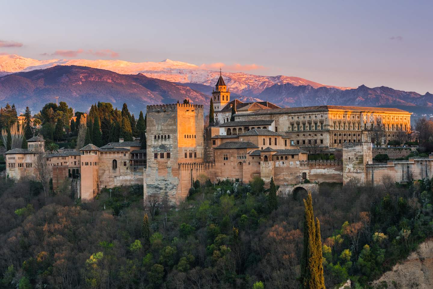 A picturesque medieval castle complex nestled among lush greenery, with majestic snow-capped mountains in the background, bathed in the warm glow of the setting sun.