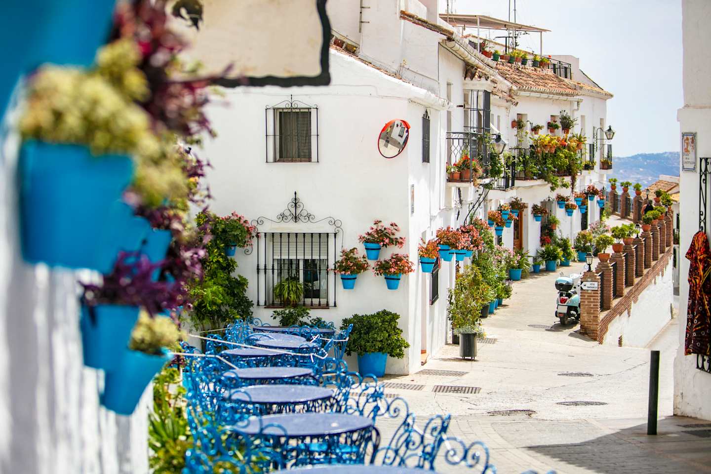 A charming, colorful alleyway lined with potted plants, flower-adorned buildings, and blue outdoor seating, creating a picturesque Mediterranean scene.