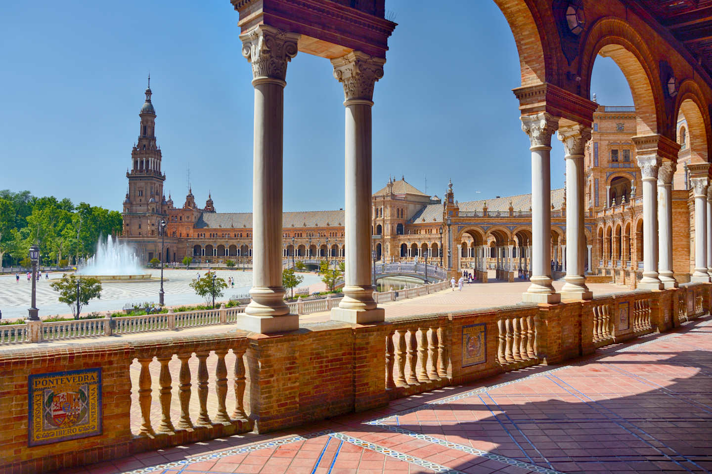 A grand, ornate architectural structure with arched columns and intricate details, set against a clear blue sky, with a central fountain and paved walkways in the foreground.