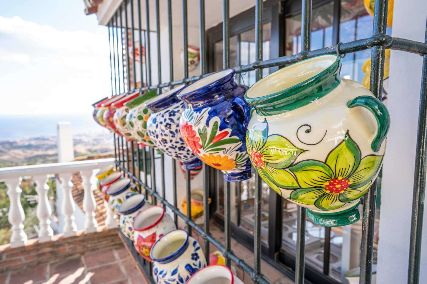 Colorful ceramic pots with floral designs are displayed on a balcony railing, overlooking a cityscape in the background.