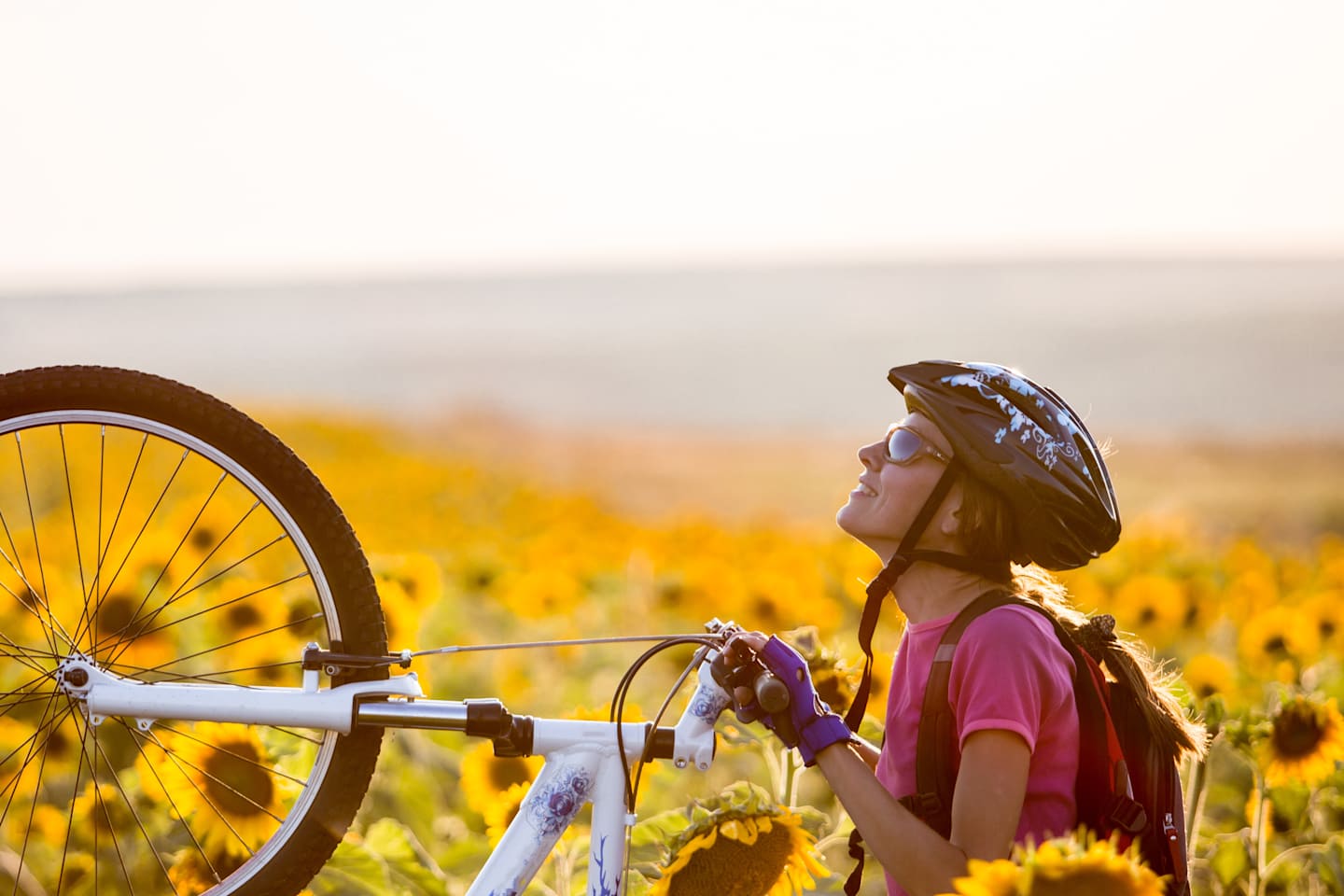 A young person in a pink shirt and cap stands next to a bicycle in a field of sunflowers, with a hazy, distant landscape in the background.