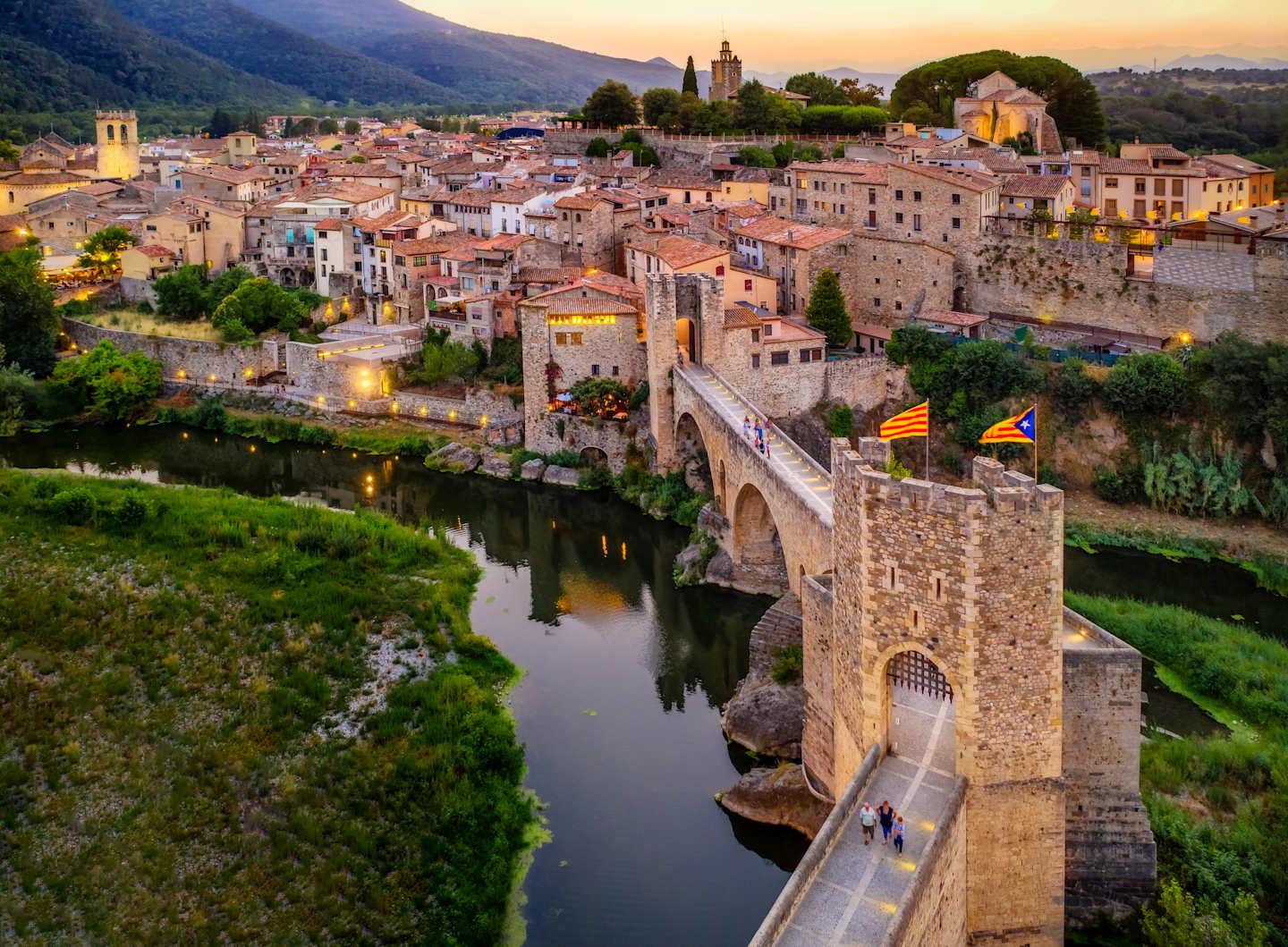 A picturesque medieval town nestled in a valley, with a historic stone bridge spanning a tranquil river, surrounded by lush greenery and a stunning mountainous backdrop at sunset.