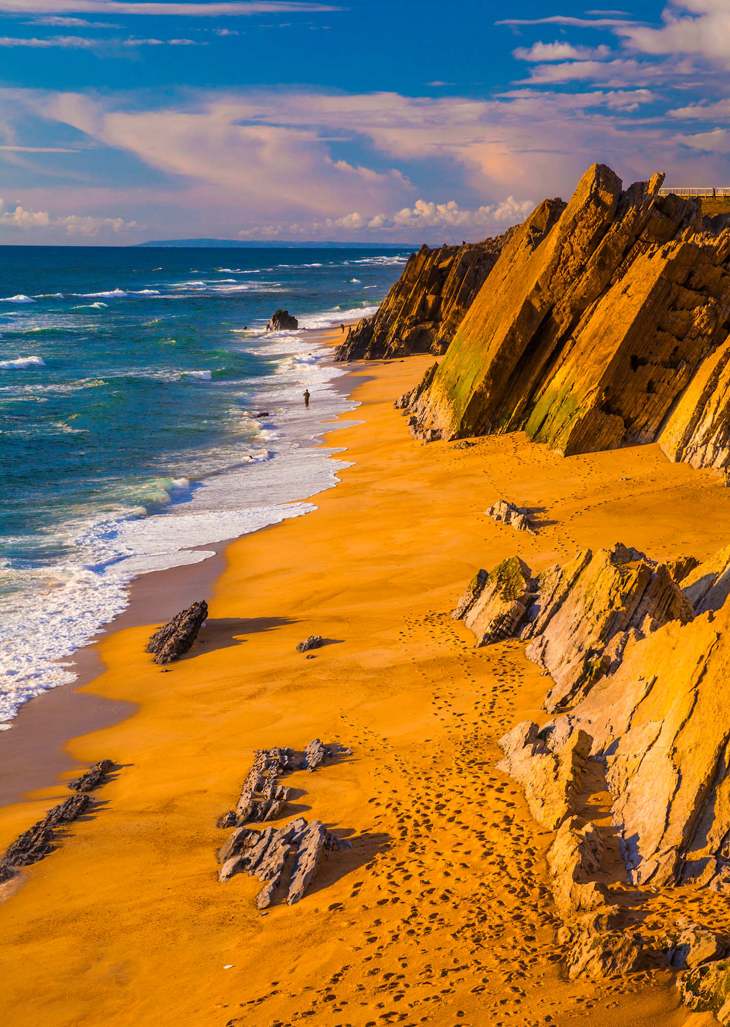 A stunning coastal landscape with rugged, golden-hued cliffs overlooking a vast, turquoise ocean under a vibrant, cloud-filled sky.