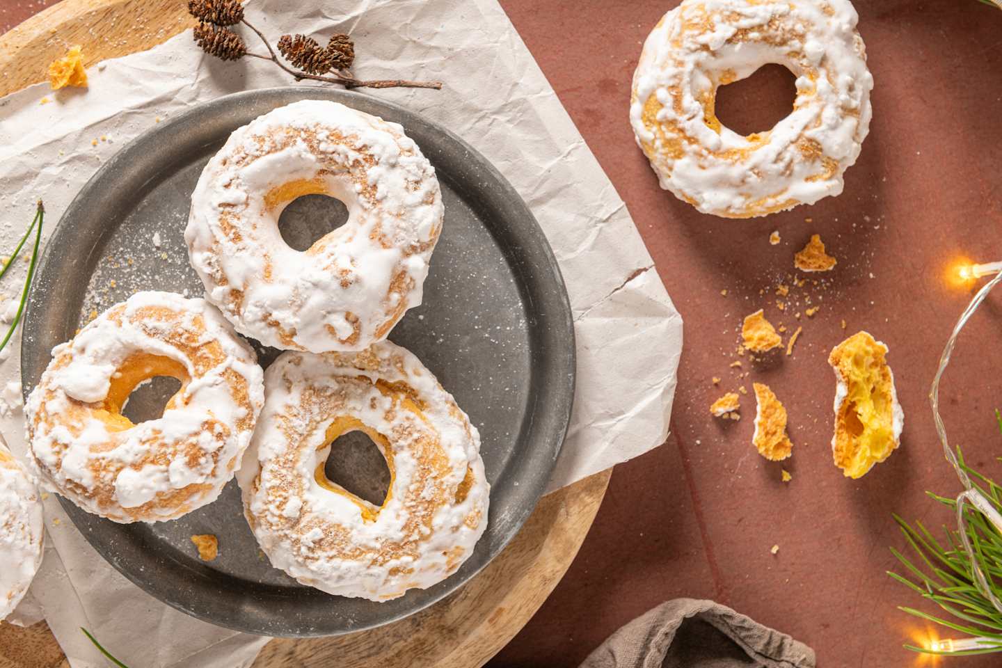 The image shows a plate of powdered sugar-coated pastries, accompanied by pine cones and greenery, set against a rustic background.