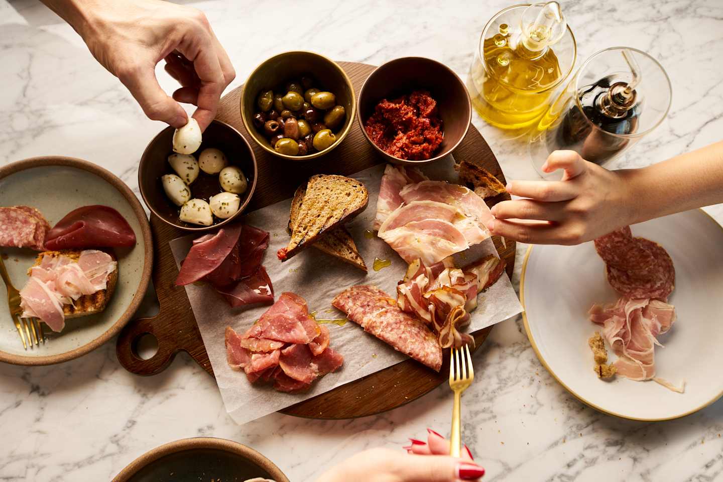 A wooden board on a marble surface displays an assortment of appetizing items, including cured meats, olives, bread, and a bottle of olive oil, all arranged in an inviting manner.